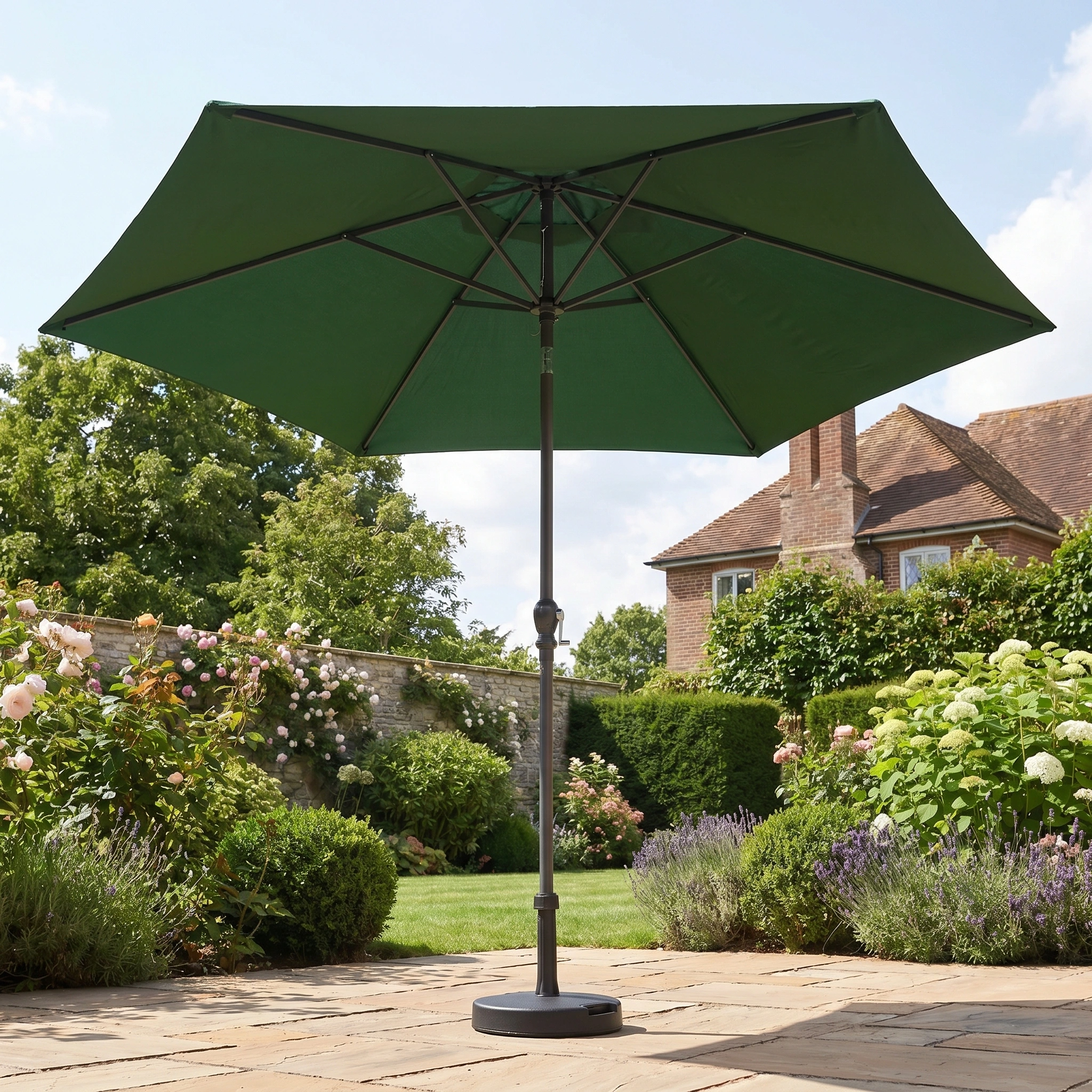 The Triton 2.5m Round Aluminium Parasol in Green stands on a stone terrace in a flower-filled garden, with a brick house visible in the background.