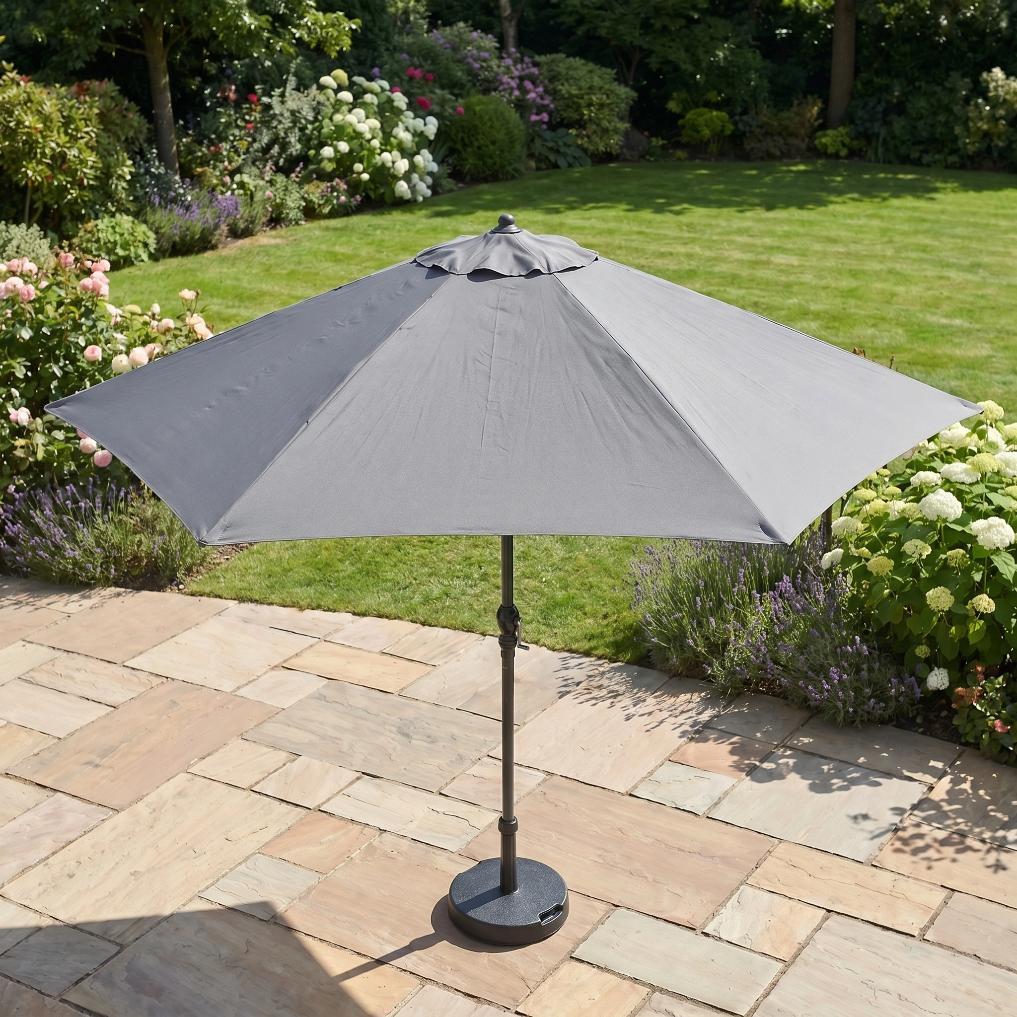 The Triton 2.5m Round Aluminium Parasol in Grey adds style and UV protection to your outdoor space, perfect for a stone patio with a green lawn and flower garden backdrop.