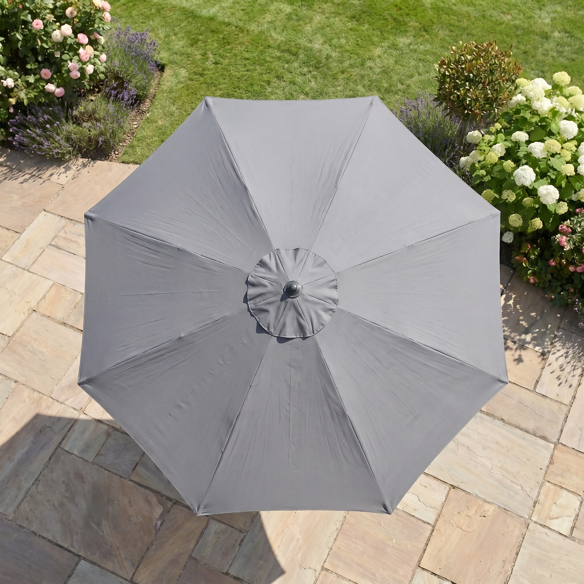 The Triton 2.5m Round Aluminium Parasol in Grey sits on a stone patio, surrounded by garden greenery—offering stylish charm and UV protection for your outdoor space.