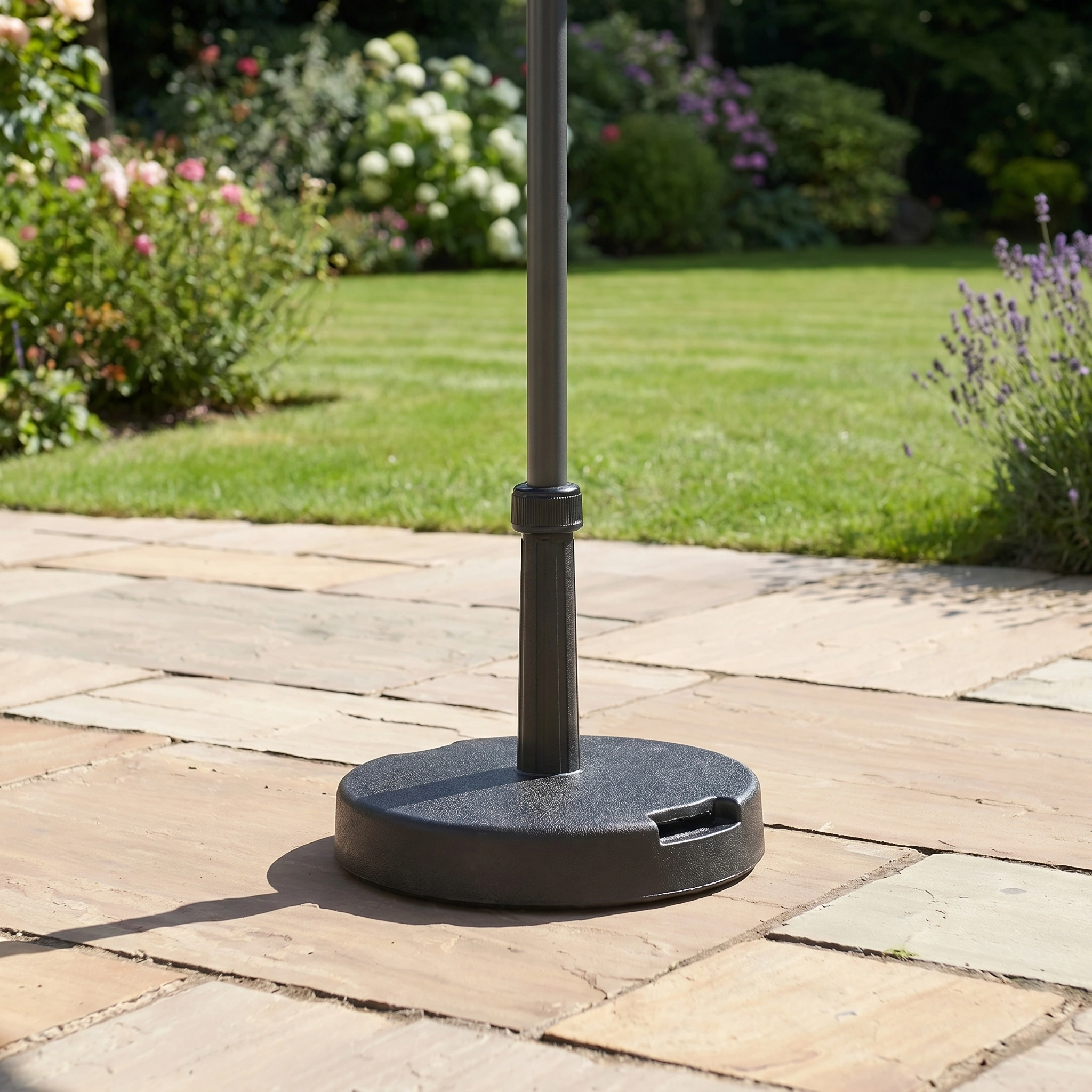 Close-up of a black stand supporting the Triton 2.5m Round Aluminium Parasol in Grey on a stone patio, with a lush green garden in the background.