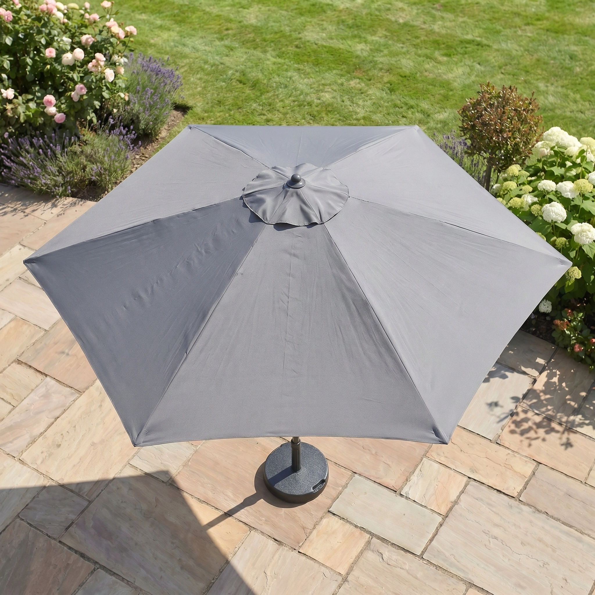 The Triton 2.5m Round Aluminium Parasol in Grey stands open on a stone patio amid grass and flowering plants, providing excellent UV protection for your garden.