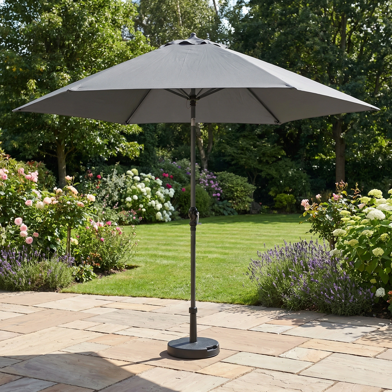 The Triton 2.5m Round Aluminium Parasol in Grey brings shade and style to your garden, standing elegantly on a stone terrace amid green grass and blooming flowers.