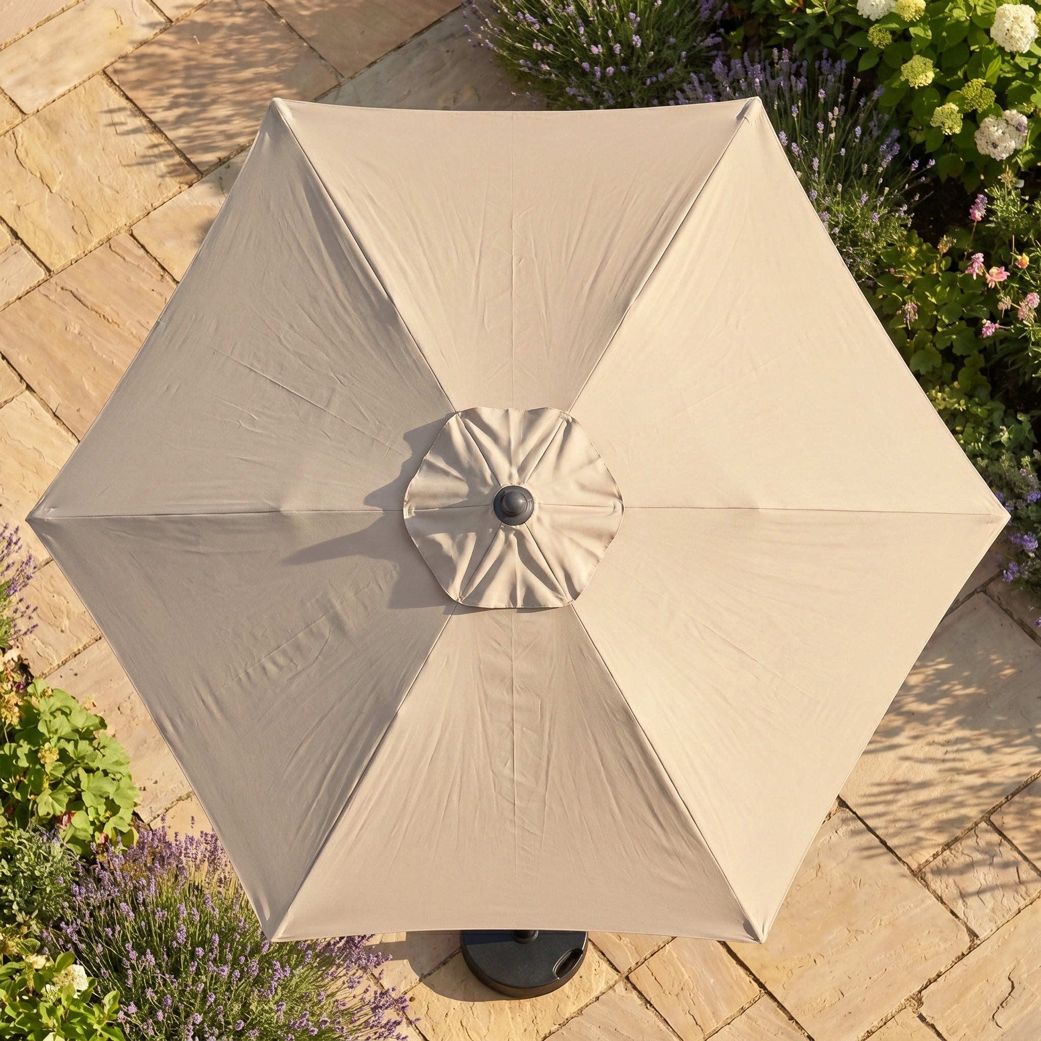 The Triton 2.5m Round Aluminium Parasol in Khaki offers UV protection and fade resistance, shown from above among stone tiles, green plants, and flowers.