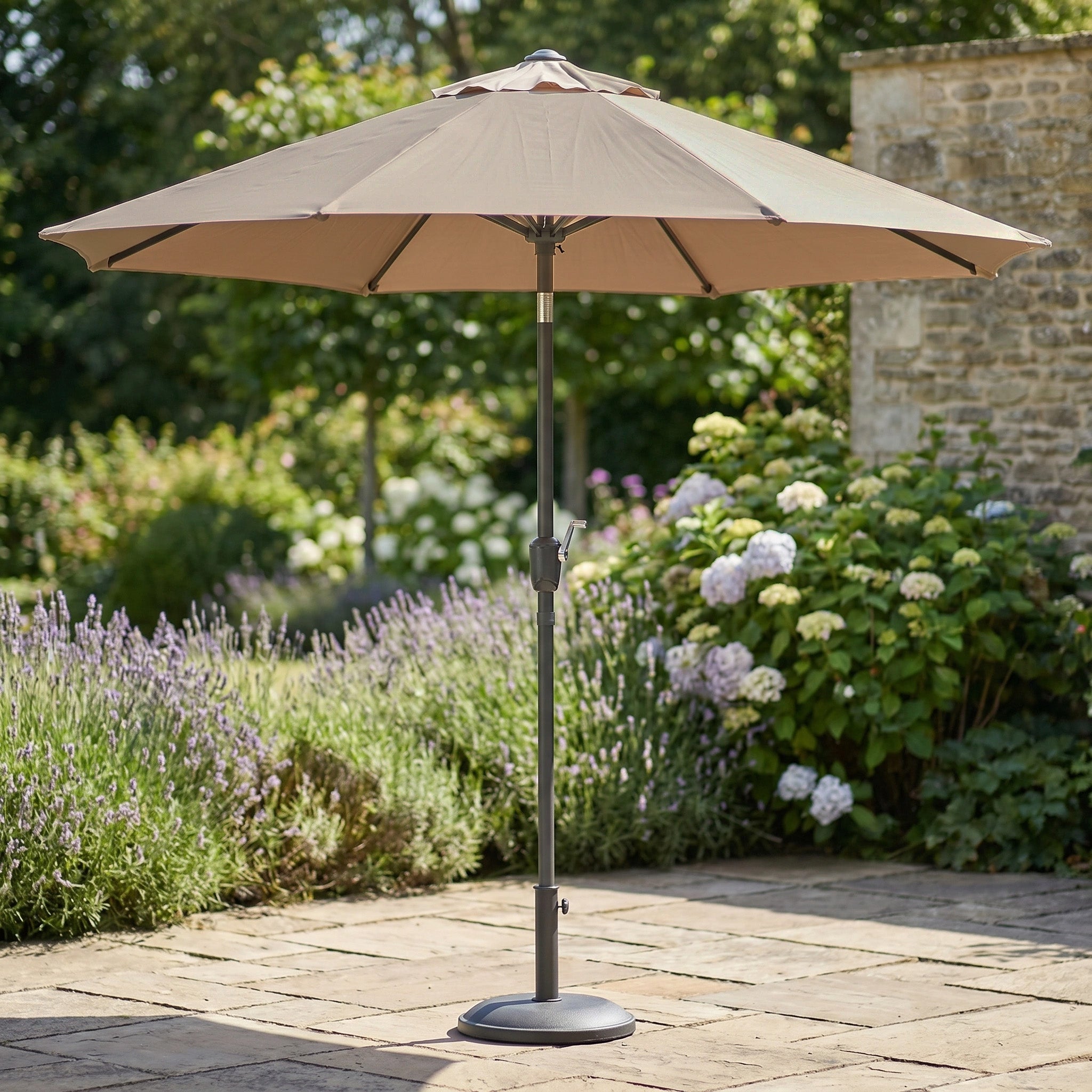The Triton 2.5m Round Aluminium Parasol in Khaki sits on a stone terrace, surrounded by blooming garden flowers—a stylish, fade-resistant parasol for relaxing outdoors.