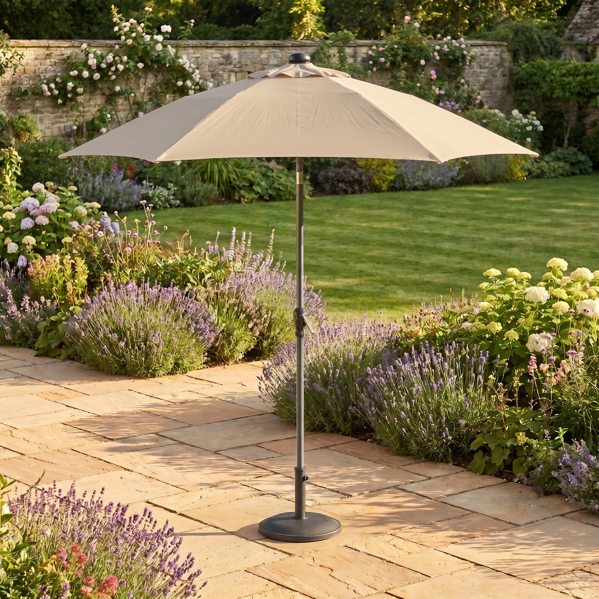 A Triton 2.5m Round Aluminium Parasol in Khaki, known for its fade resistance, stands on a stone terrace in a garden filled with blooming flowers and green grass.
