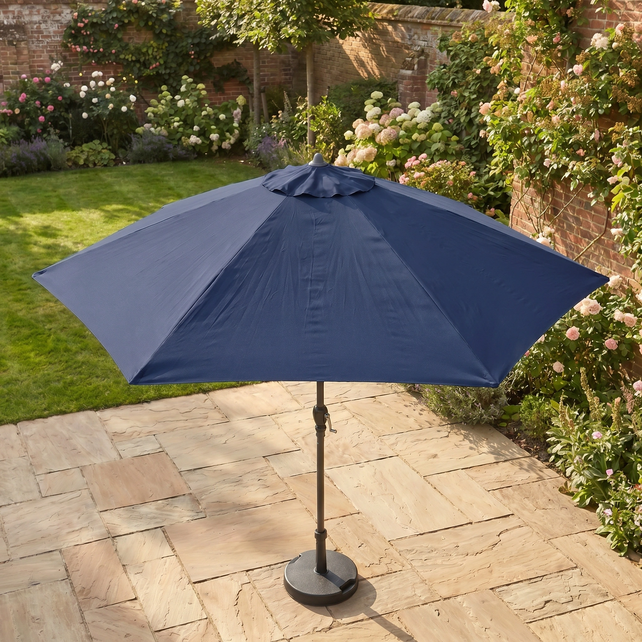 The Triton 2.5m Round Aluminium Parasol in Navy with UV protection stands on a stone terrace in a garden full of flowers and greenery.