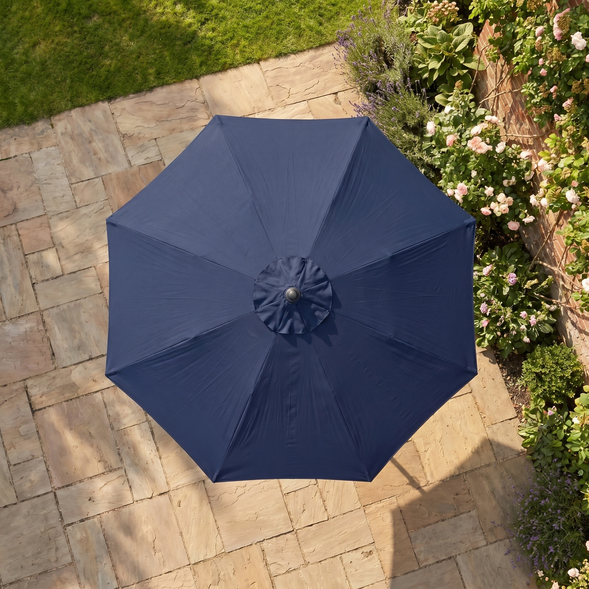 The Triton 2.5m Round Aluminium Parasol in Navy with UV protection is shown from above on a stone patio beside a grassy garden filled with blooming flowers.