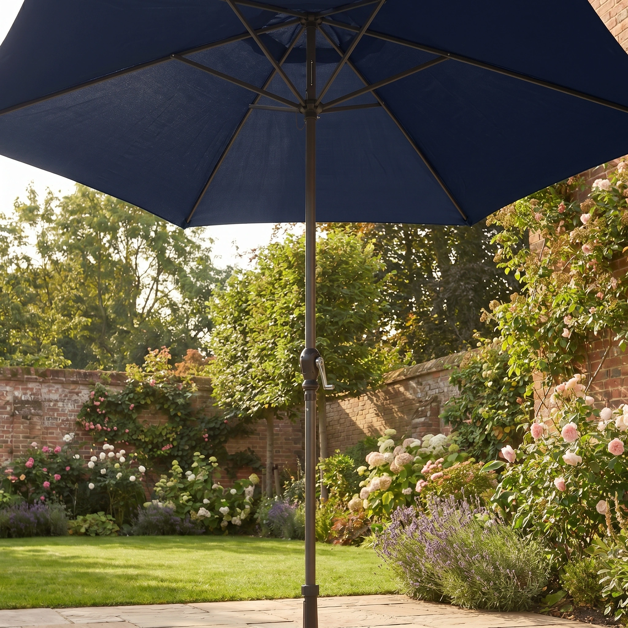The Triton 2.5m Round Aluminium Parasol in Navy, featuring UV protection, stands in a sunny garden filled with flowers and bordered by a brick wall.