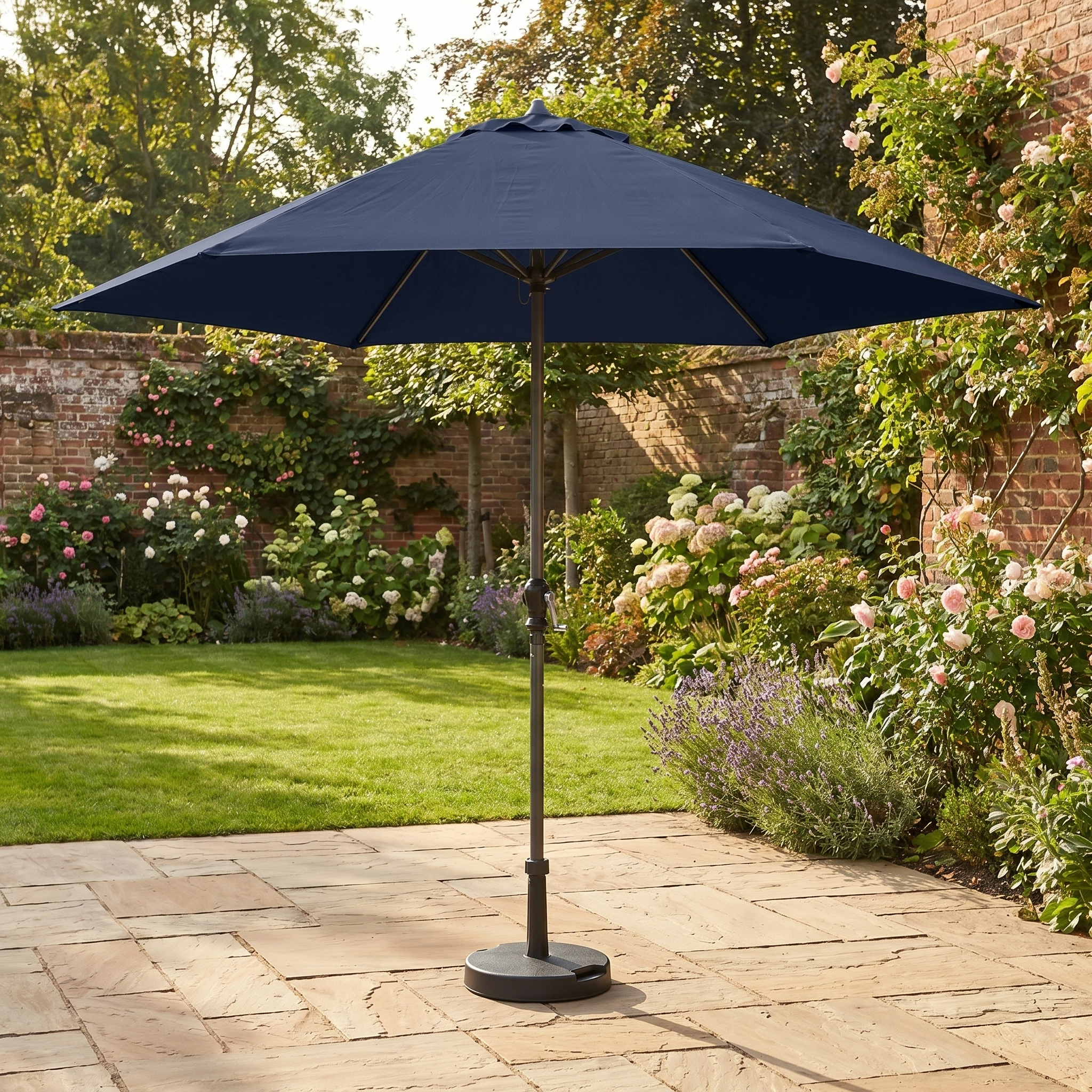 The Triton 2.5m Round Aluminium Parasol in Navy stands on a stone patio in a sunny, flower-filled garden, providing excellent UV protection.