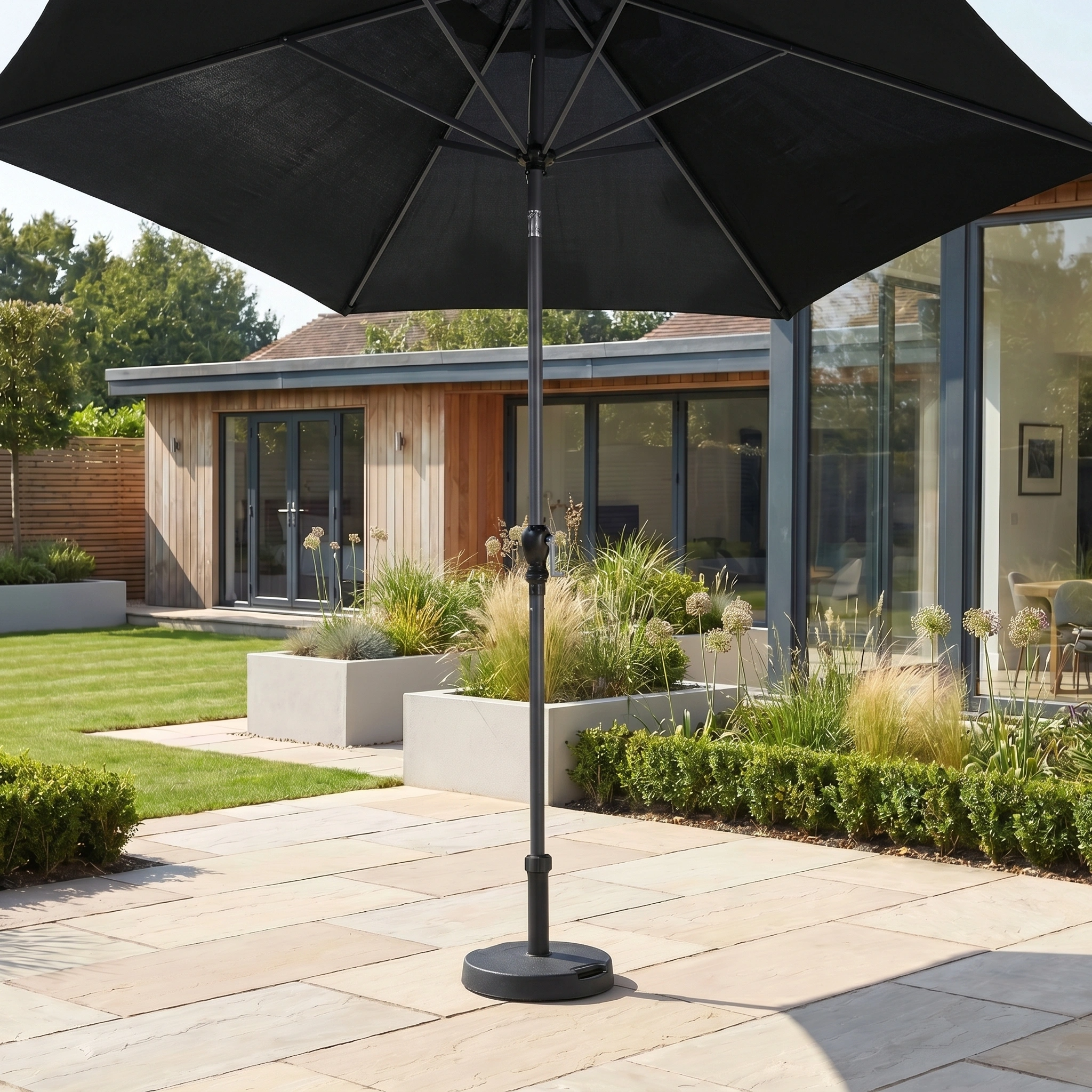 The Triton 3m Round Aluminium Parasol in Black provides shade in a sunny modern backyard, surrounded by greenery and a contemporary house in the background.
