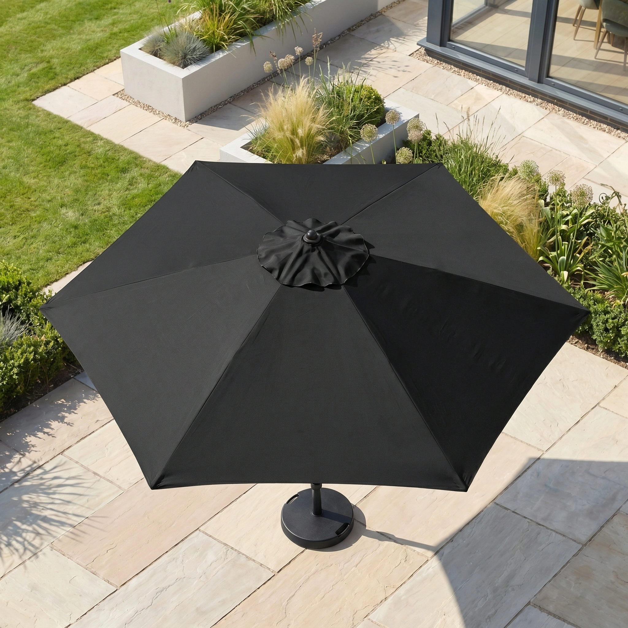 The Triton 3m Round Aluminium Parasol in Black with a crank handle stands on a stone patio amid modern landscaping and green lawn, providing UV protection for outdoor comfort.