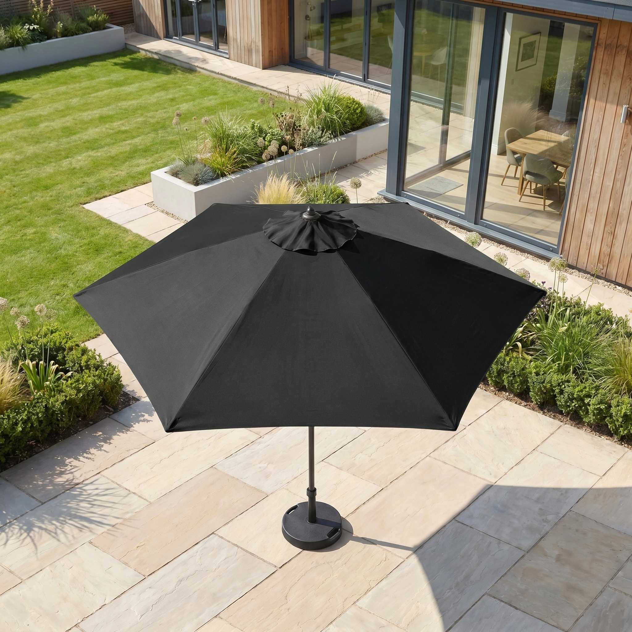 The Triton 3m Round Aluminium Parasol in Black stands on a stone terrace beside a house with glass doors, providing stylish UV protection in the modern garden.
