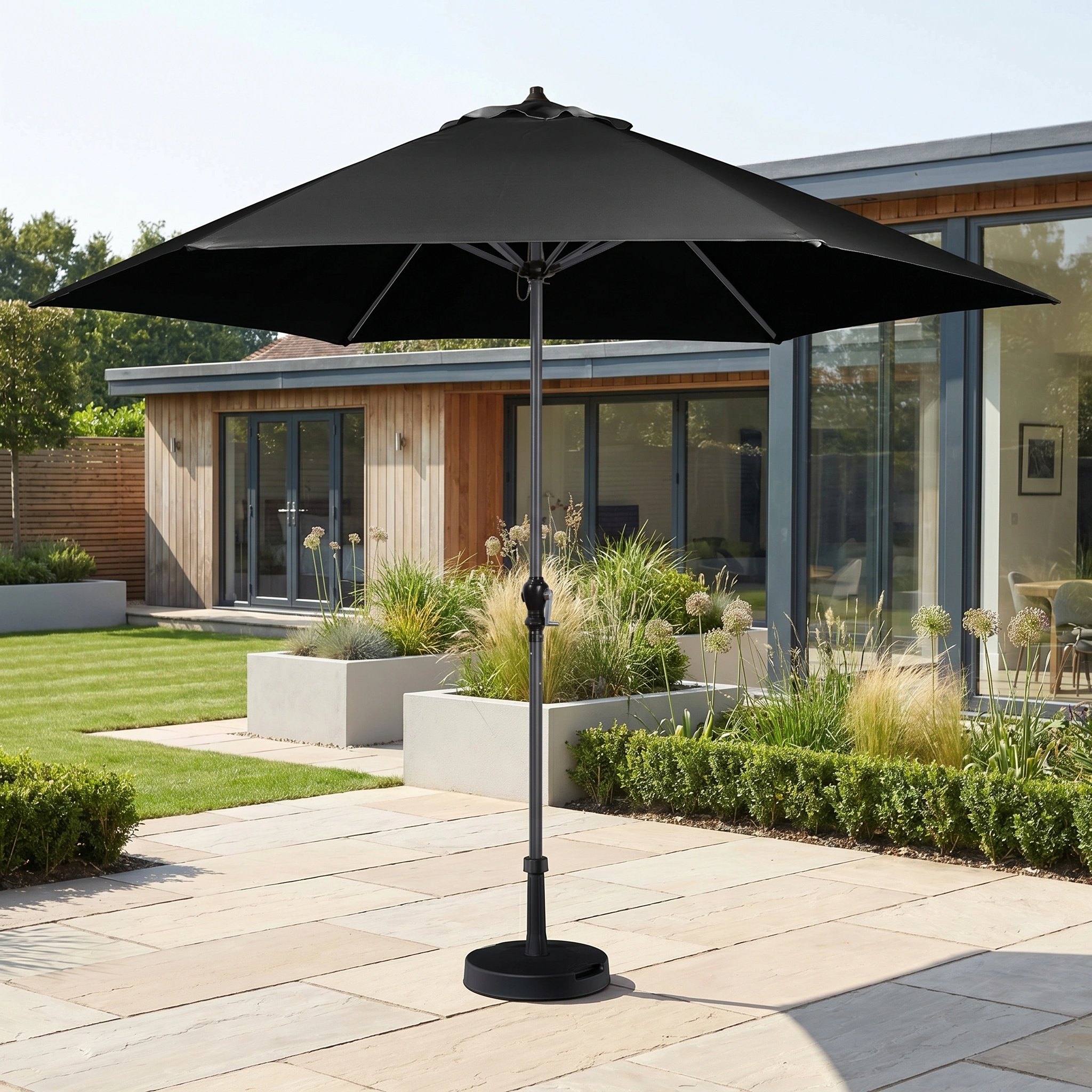 The Triton 3m Round Aluminium Parasol in Black stands open on a stone terrace beside a house, providing stylish shade and UV protection in a modern garden.