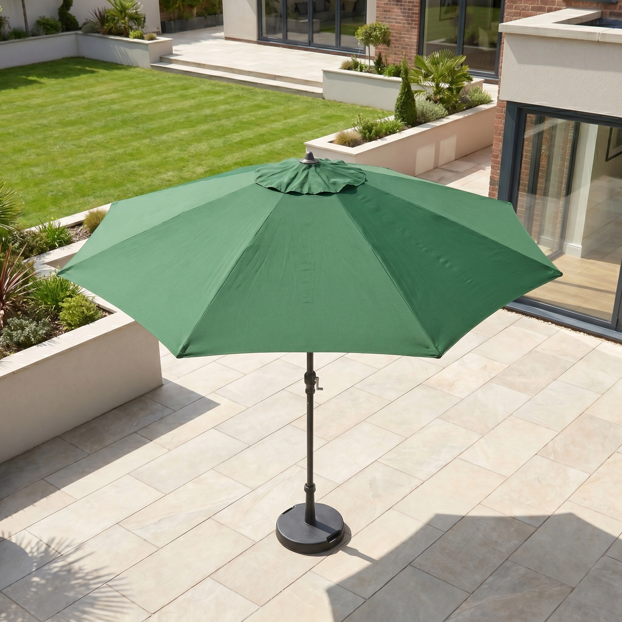 The Triton 3m Round Aluminium Parasol in Green provides UV protection to your tiled outdoor area, with a lawn and modern house in the background—perfect for relaxing outdoors.