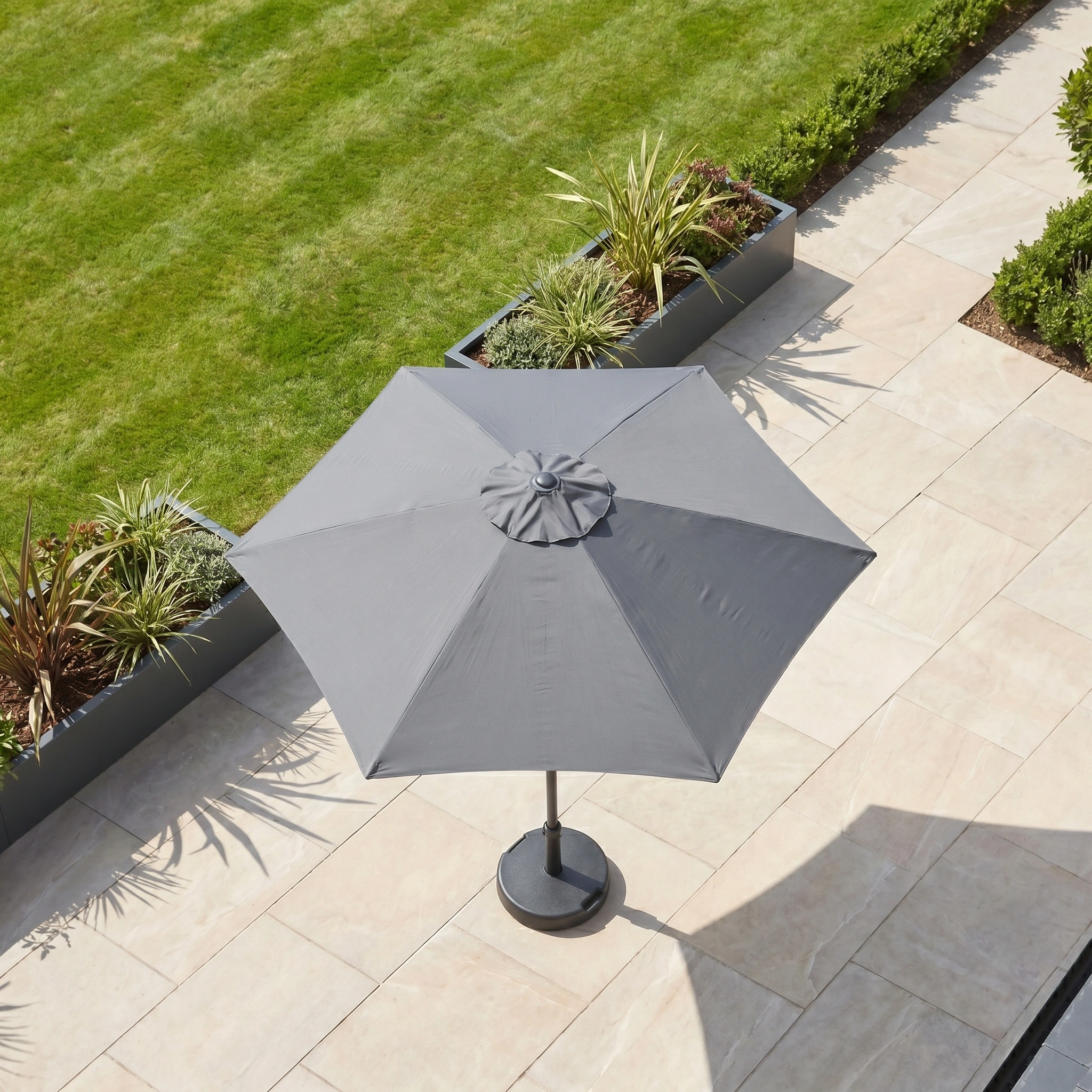 The Triton 3m Round Aluminium Parasol in Grey is on a tiled terrace near green grass and garden planters, viewed from above.