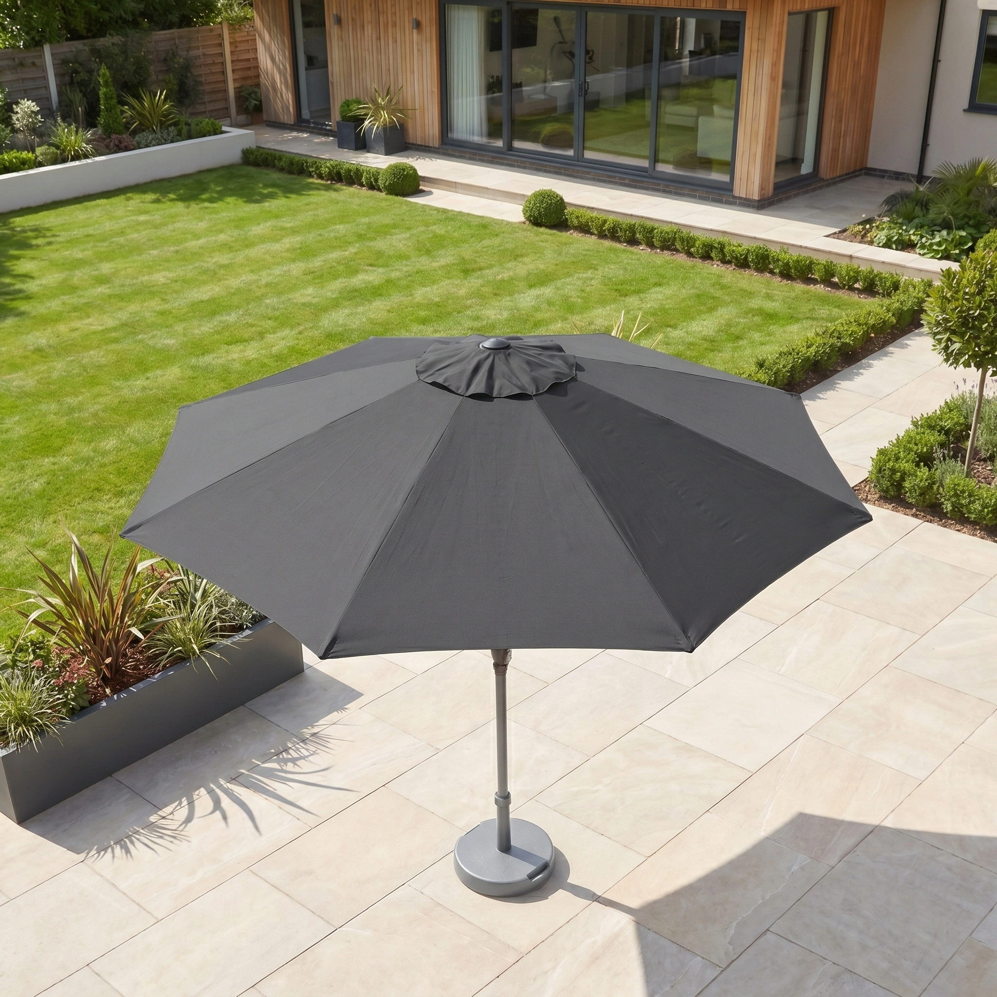 The Triton 3m Round Aluminium Parasol in Grey stands on a tiled terrace beside a modern house with a green lawn and garden, offering stylish shade and UV protection.