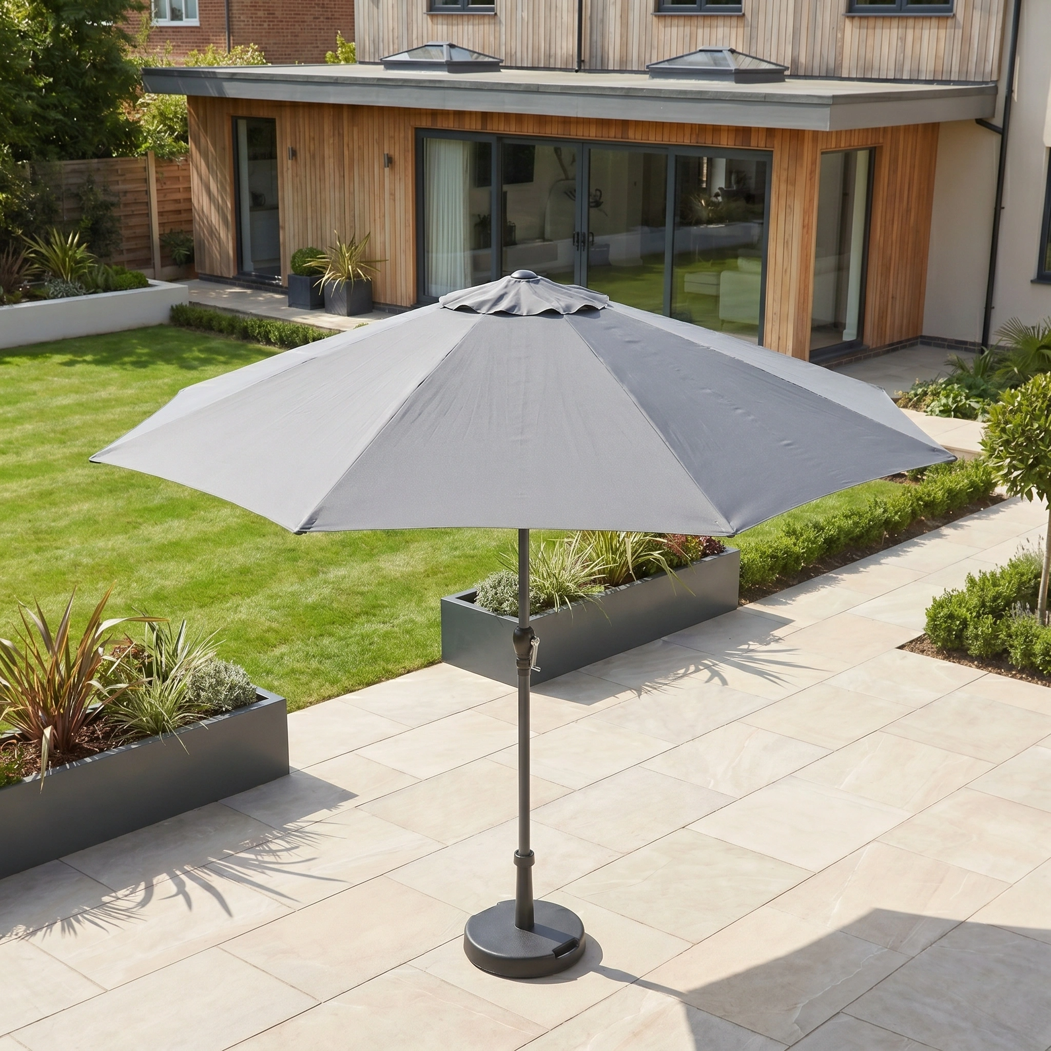 The Triton 3m Round Aluminium Parasol in Grey on a tiled terrace with green lawn and modern house in the background provides stylish shade and UV protection.