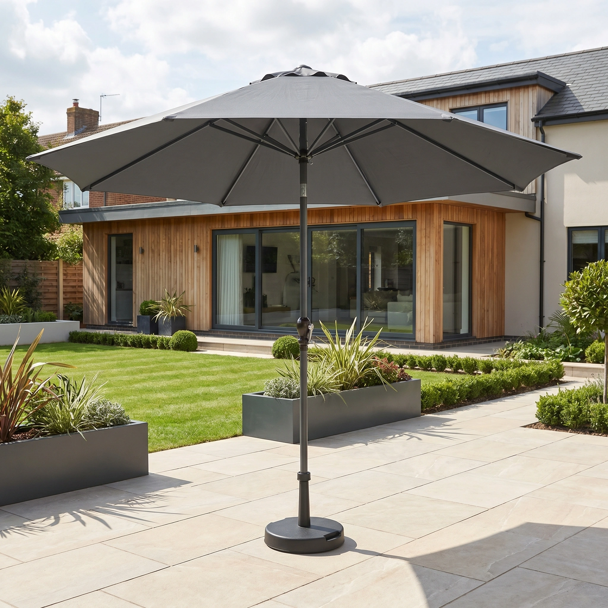 The Triton 3m Round Aluminium Parasol in Grey stands on a tiled terrace before a modern house with a lush garden, providing stylish shade and comfort.