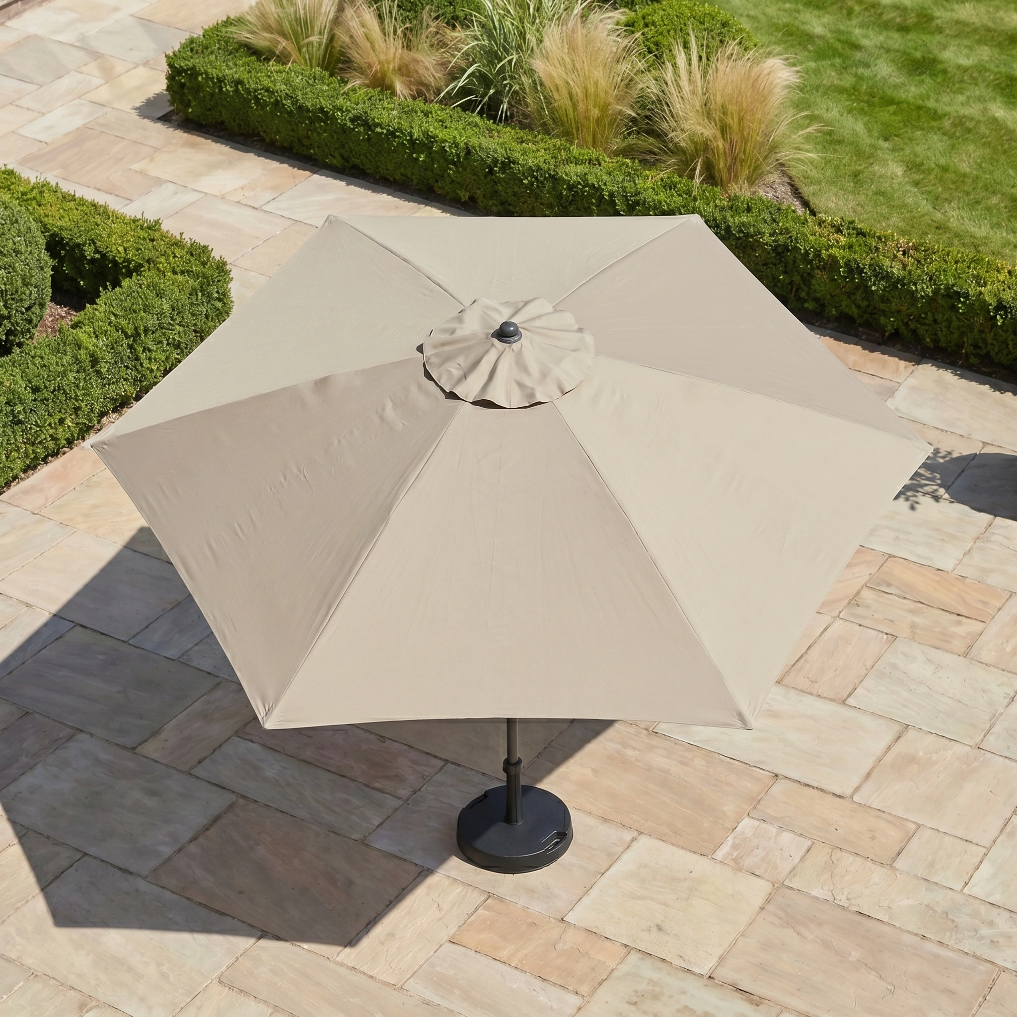 The Triton 3m Round Aluminium Parasol in Khaki stands closed on a stone patio amid manicured hedges and ornamental grasses, providing style and UV protection.