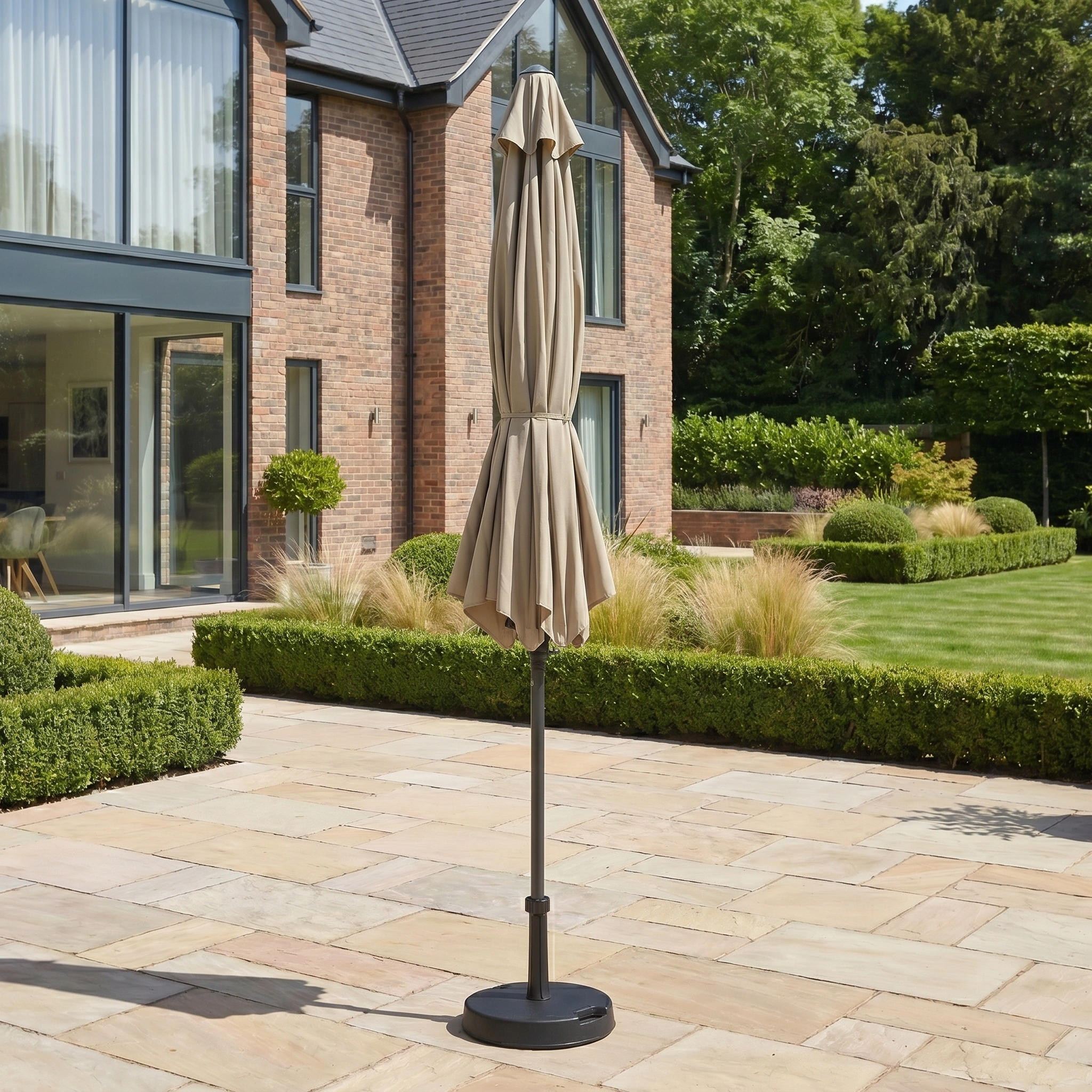 A closed Triton 3m Round Aluminium Parasol in Khaki stands on a stone terrace in a landscaped garden by a modern brick house.