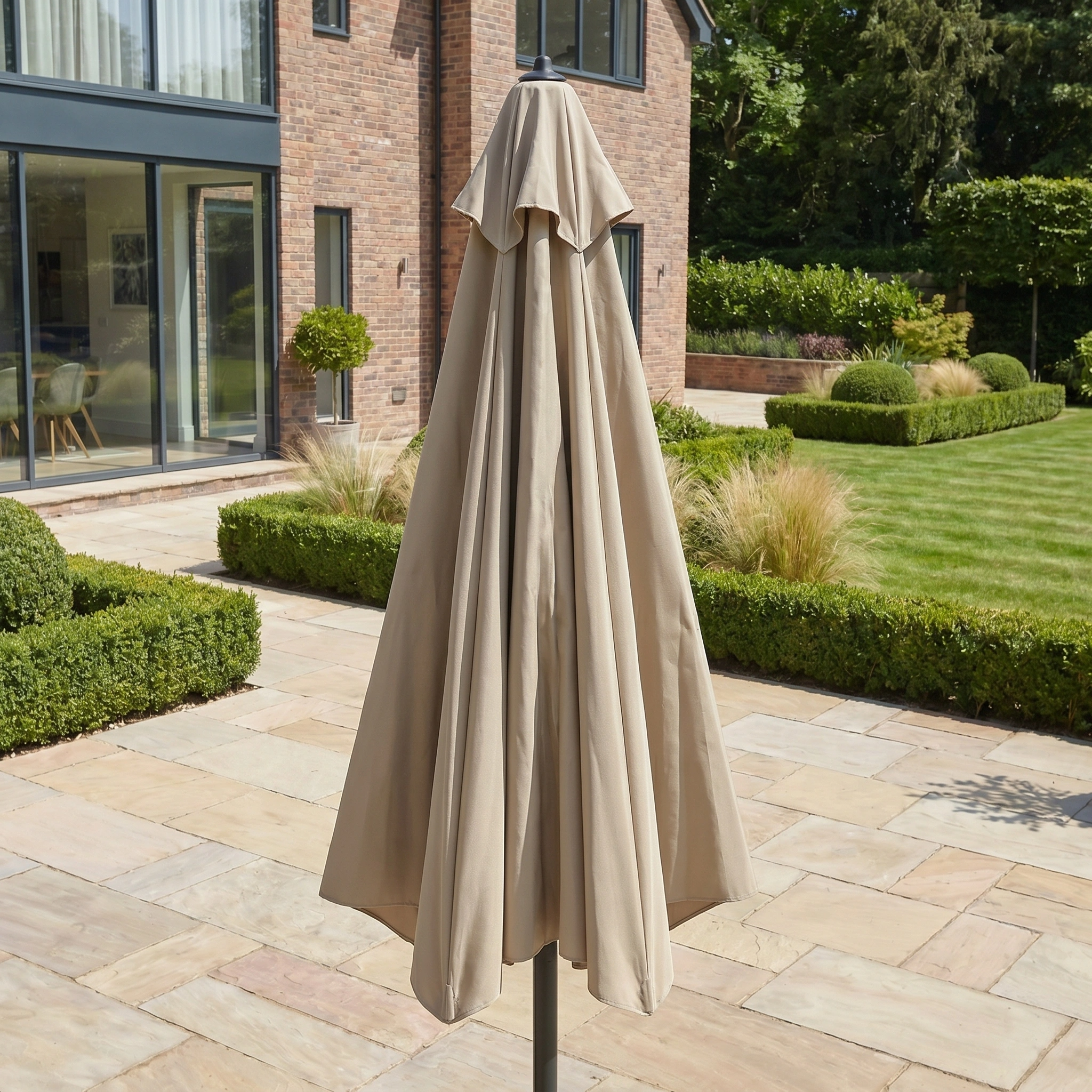 A closed Triton 3m Round Aluminium Parasol in Khaki stands on a stone terrace in a landscaped garden beside a modern brick house, ready to provide UV protection.