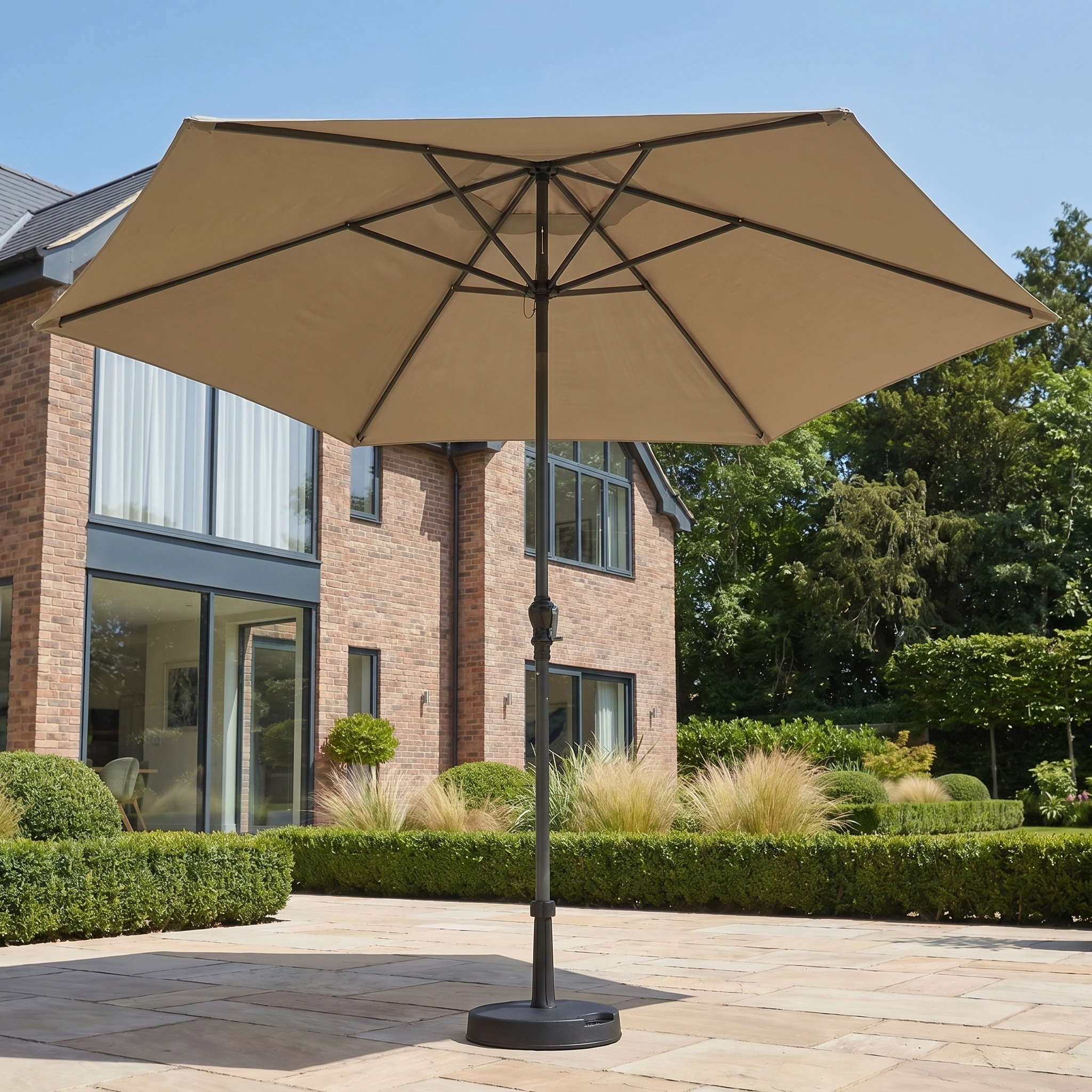 The Triton 3m Round Aluminium Parasol in Khaki features a crank handle and adds stylish UV protection to outdoor spaces, ideal for terraces or gardens beside modern brick houses with beautiful landscaping.