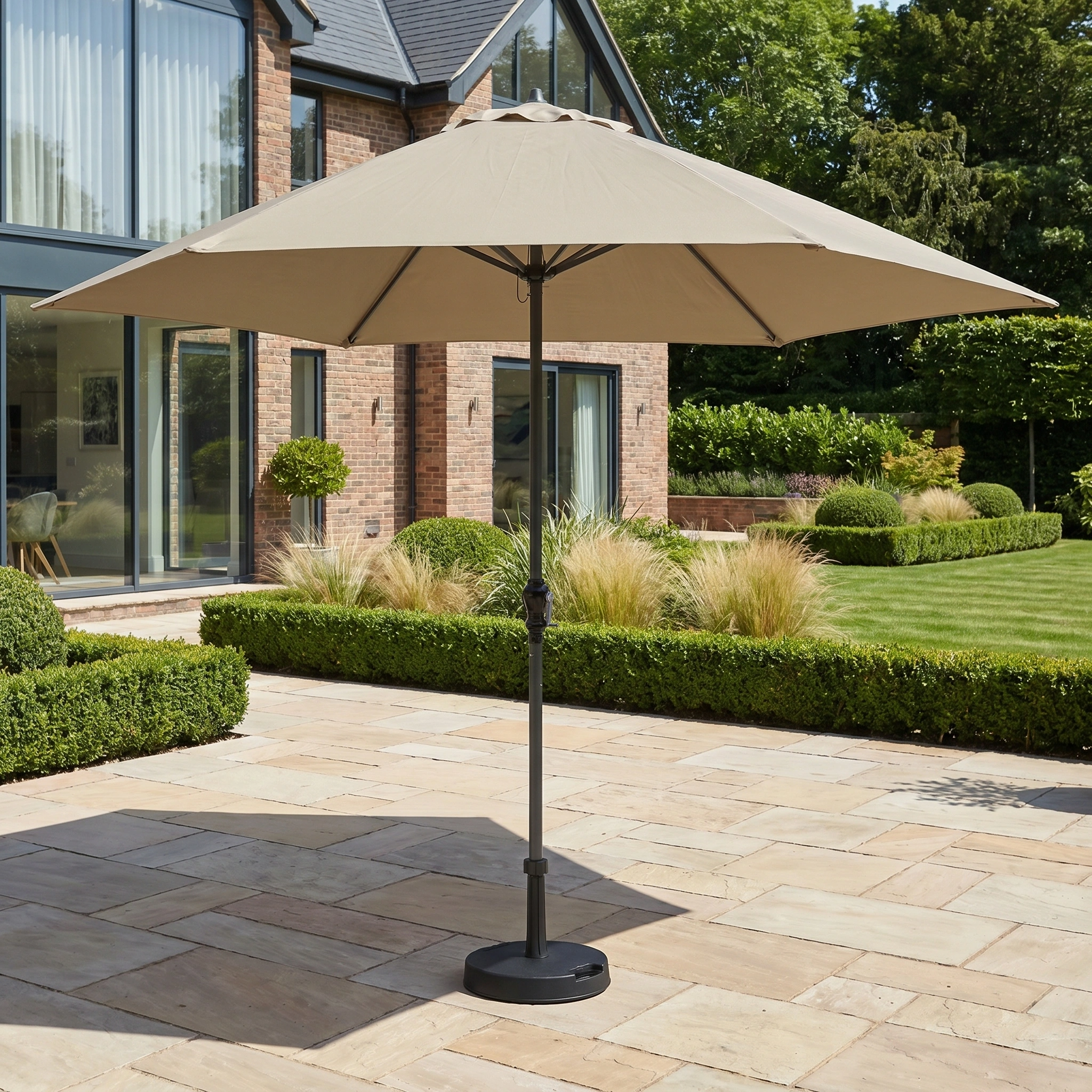 The Triton 3m Round Aluminium Parasol in Khaki shades a paved patio and manicured garden, provides UV protection, and is set against a modern house backdrop.