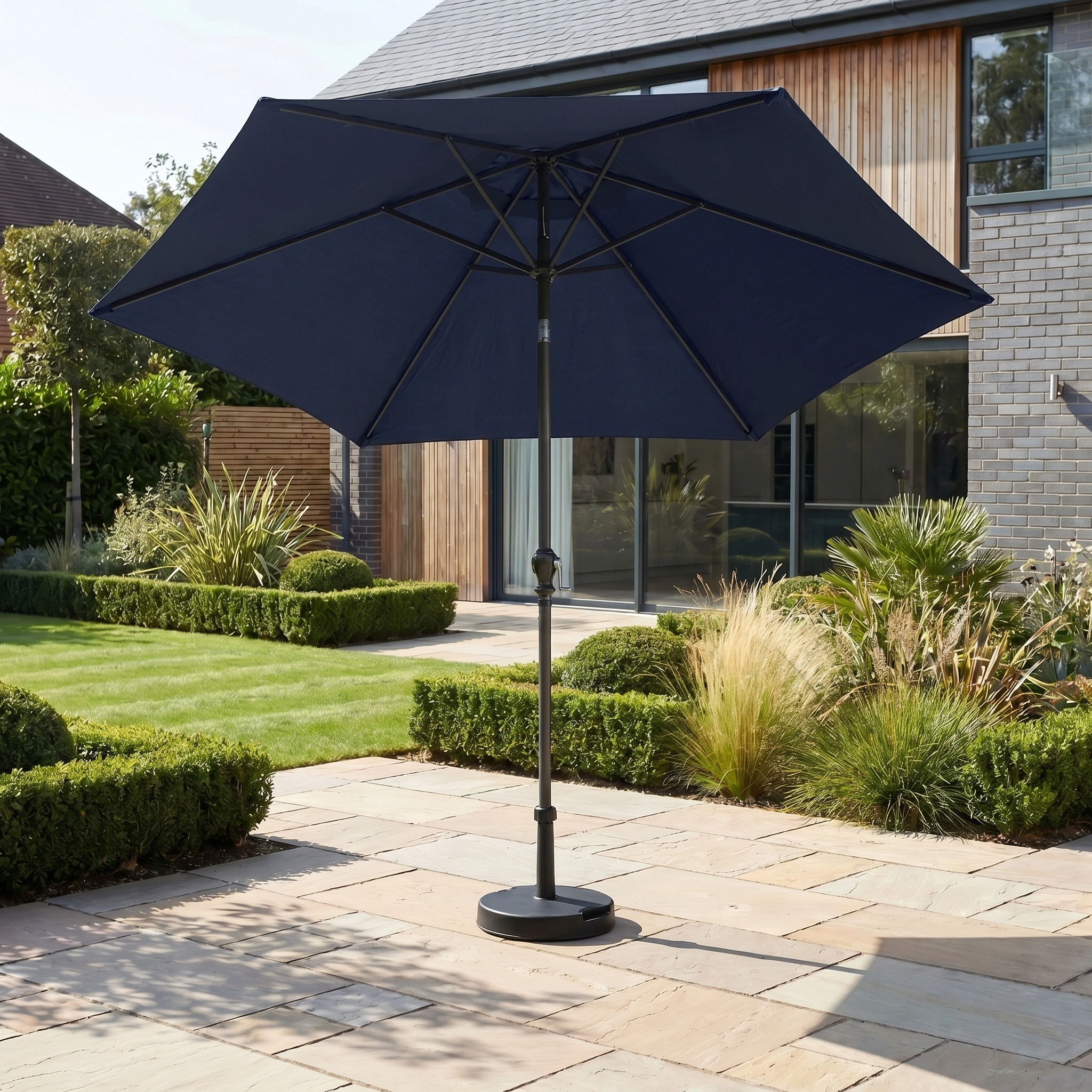 The Triton 3m Round Aluminium Parasol in Navy provides stylish shade and UV protection on a stone terrace in a modern garden beside a house.
