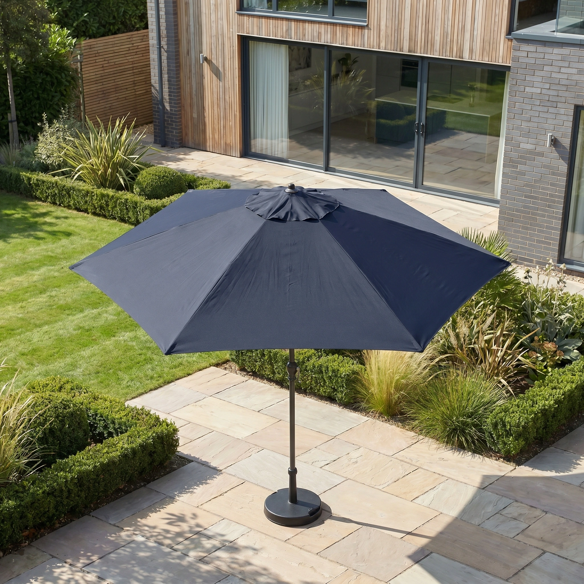 The Triton 3m Round Aluminium Parasol in Navy provides stylish shade and UV protection on a tiled terrace in a landscaped garden beside a modern house.