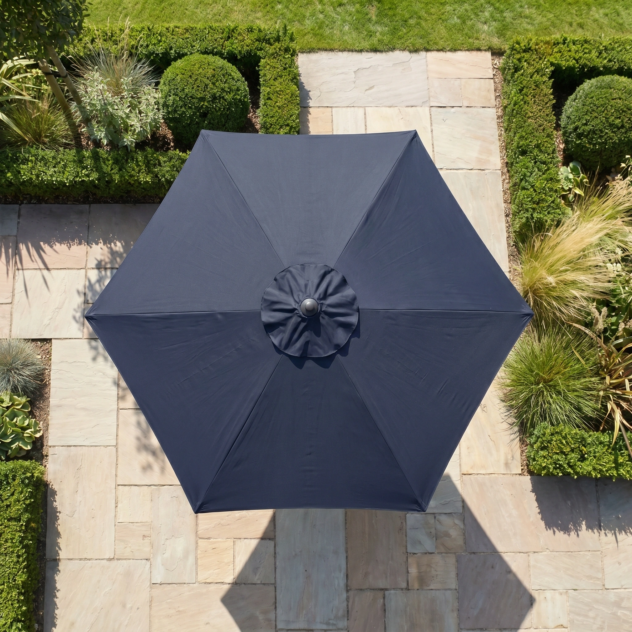 The Triton 3m Round Aluminium Parasol in Navy is shown from above, centered on stone tiles in a landscaped garden, providing stylish shade and UV protection.
