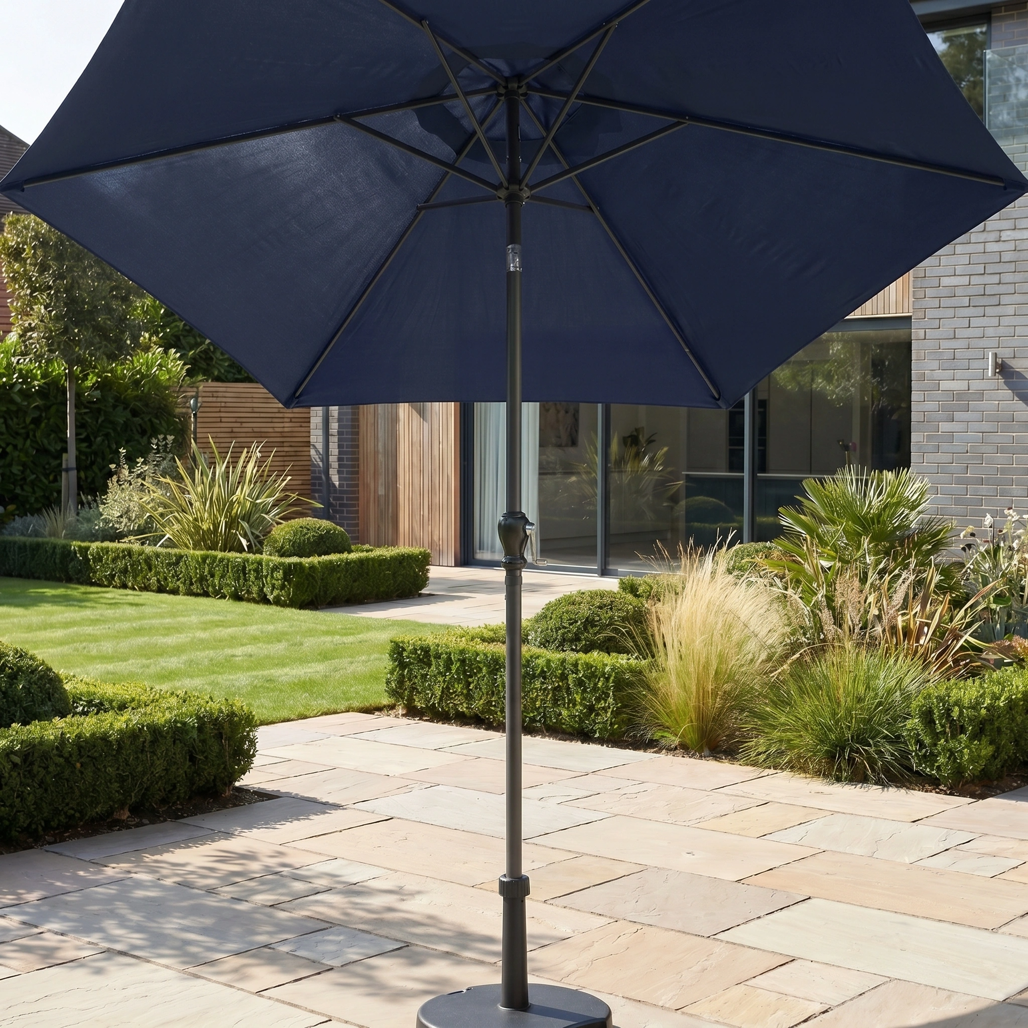 The Triton 3m Round Aluminium Parasol in Navy with UV protection stands on a stone terrace in a modern landscaped garden.