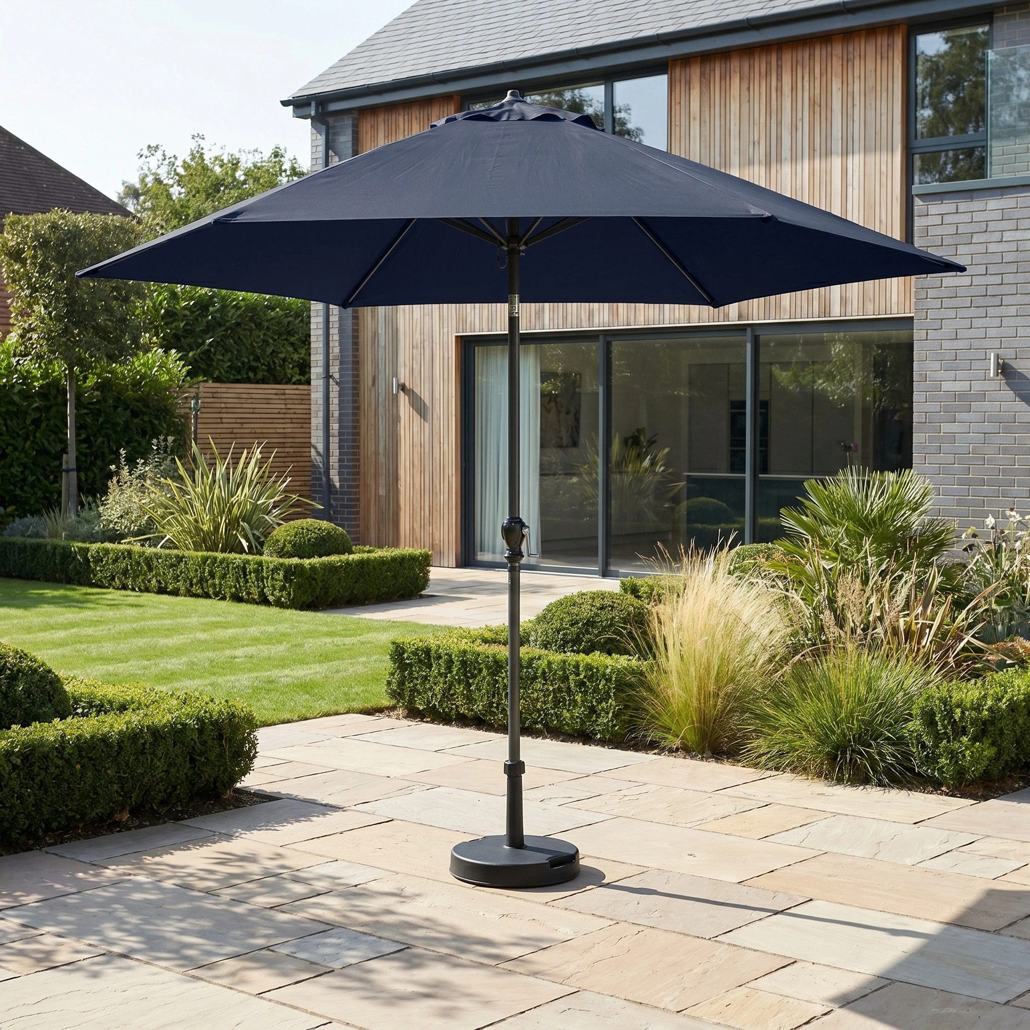 The Triton 3m Round Aluminium Parasol in Navy stands on a stone patio in a modern landscaped garden, adding style and providing UV protection.