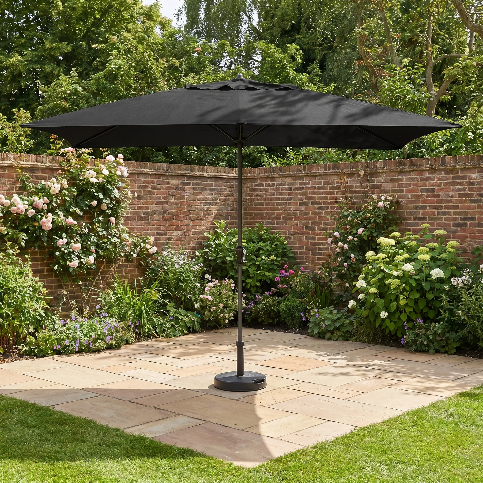 The Triton 3m x 2m Rectangular Aluminium Parasol in Black offers UV protection and stylish shade on paving stones in a flower-filled garden beside a brick wall.