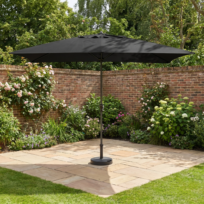 The Triton 3m x 2m Rectangular Aluminium Parasol in Black provides ample UV protection, perfect for relaxing in your garden on a paved area surrounded by flowers and a brick wall.