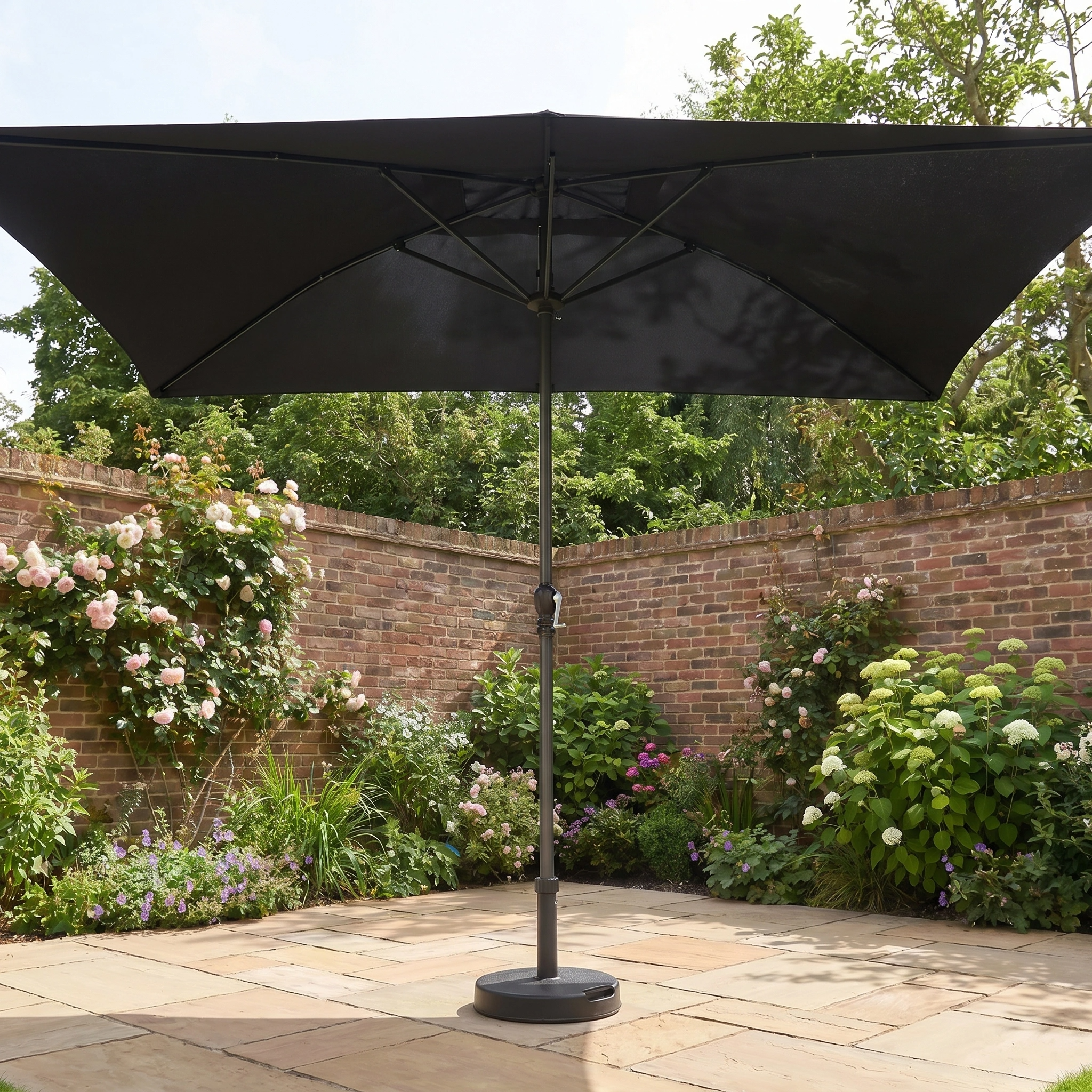 The Triton 3m x 2m Rectangular Aluminium Parasol in Black stands on a stone terrace in a walled garden with blooming flowers, providing stylish shade and UV protection.