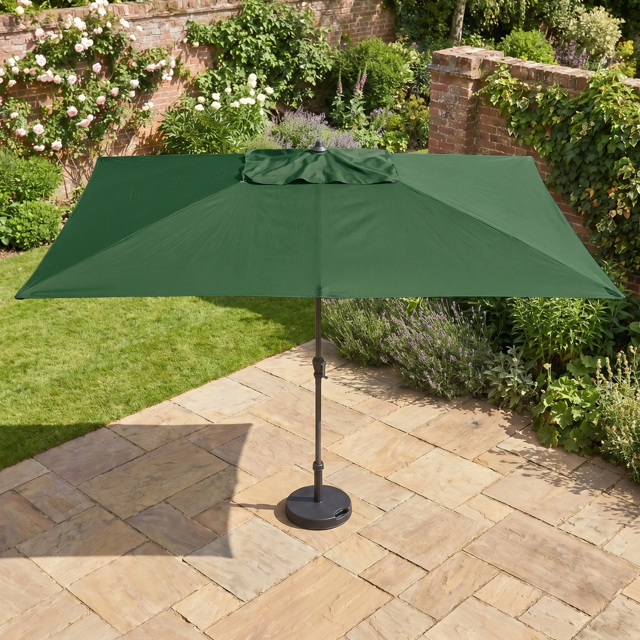 The Triton 3m x 2m Rectangular Aluminium Parasol in Green stands on a stone terrace in a garden with grass and brick walls.
