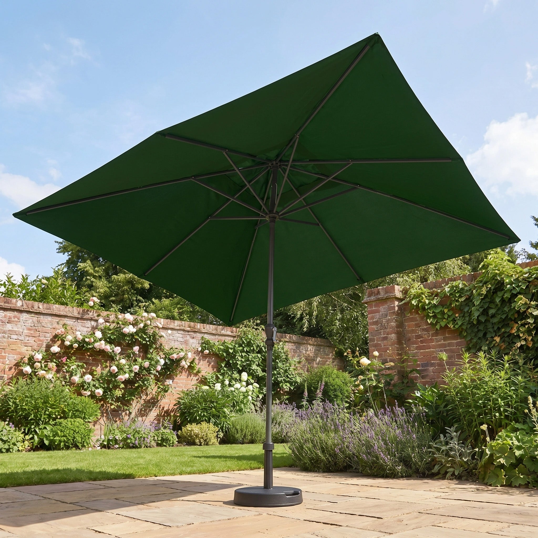 The Triton 3m x 2m Rectangular Aluminium Parasol in Green, offering UV protection, stands on a stone patio in a garden filled with flowers and lush greenery.