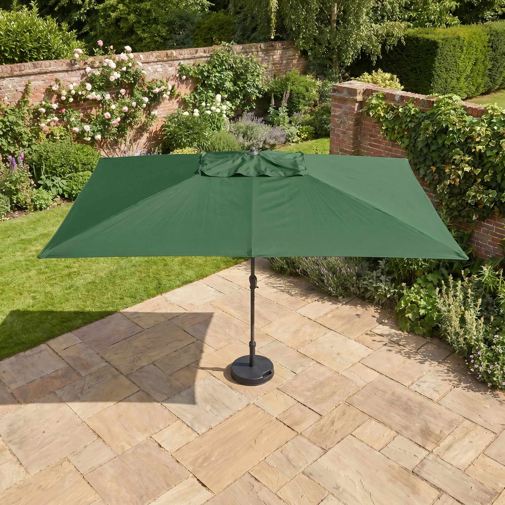 The Triton 3m x 2m Rectangular Aluminium Parasol in Green stands on a stone terrace in a sunny garden, surrounded by brick walls and lush greenery.