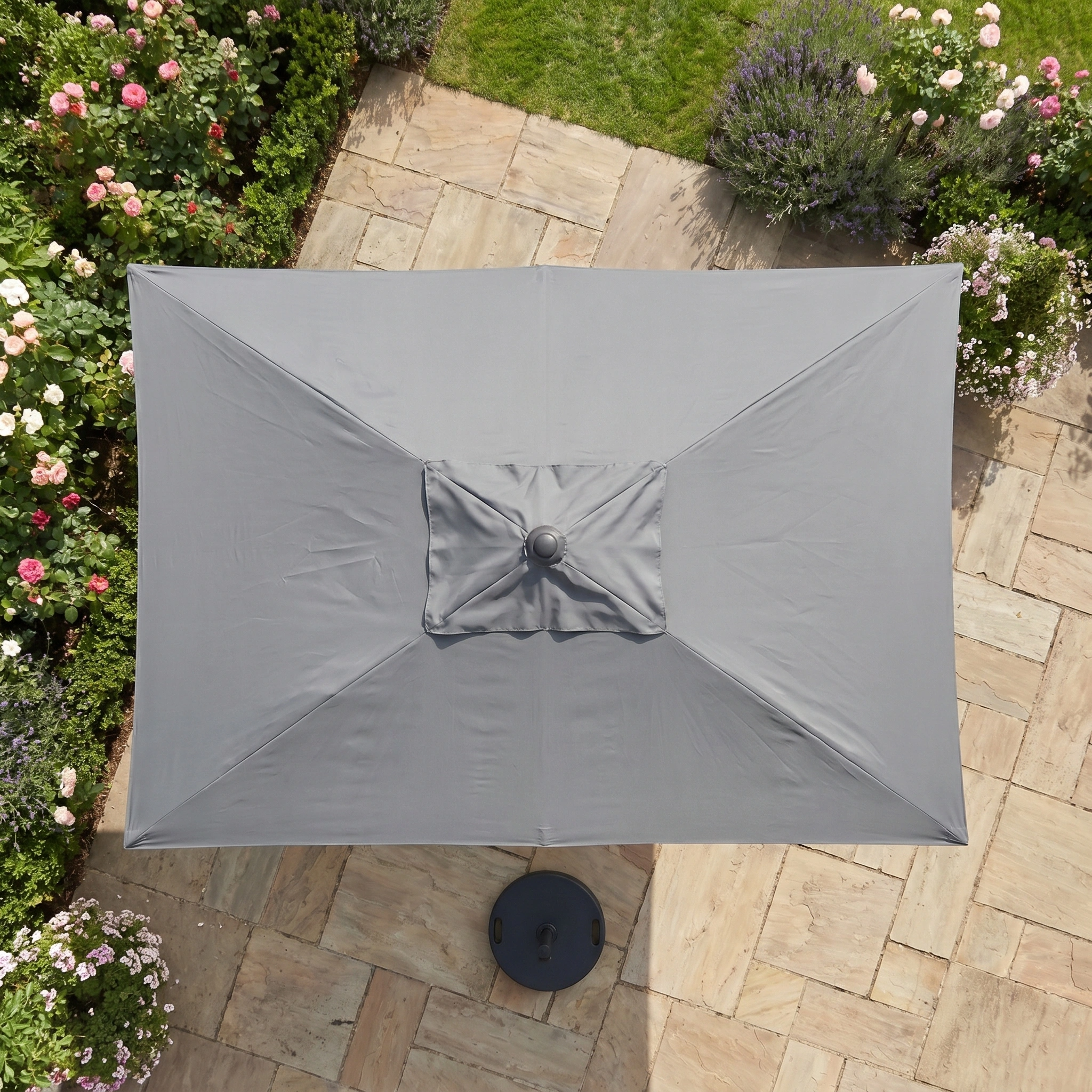 The Triton 3m x 2m Rectangular Aluminium Parasol in Grey stands on a stone patio amid blooming garden plants, providing stylish shade and reliable UV protection.