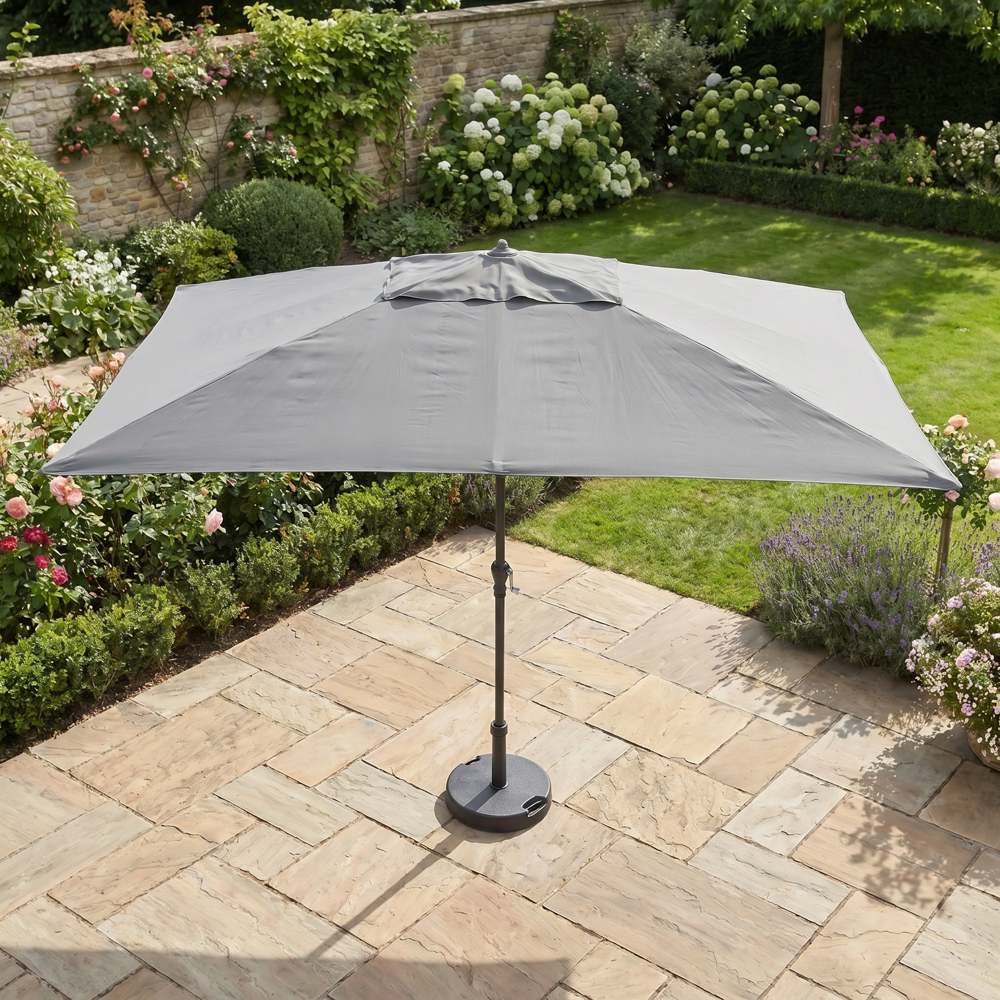 The Triton 3m x 2m Rectangular Aluminium Parasol in Grey provides UV sun protection and shade on a stone-tiled terrace, ideal for relaxing amid landscaped gardens with vibrant flowers and shrubs.