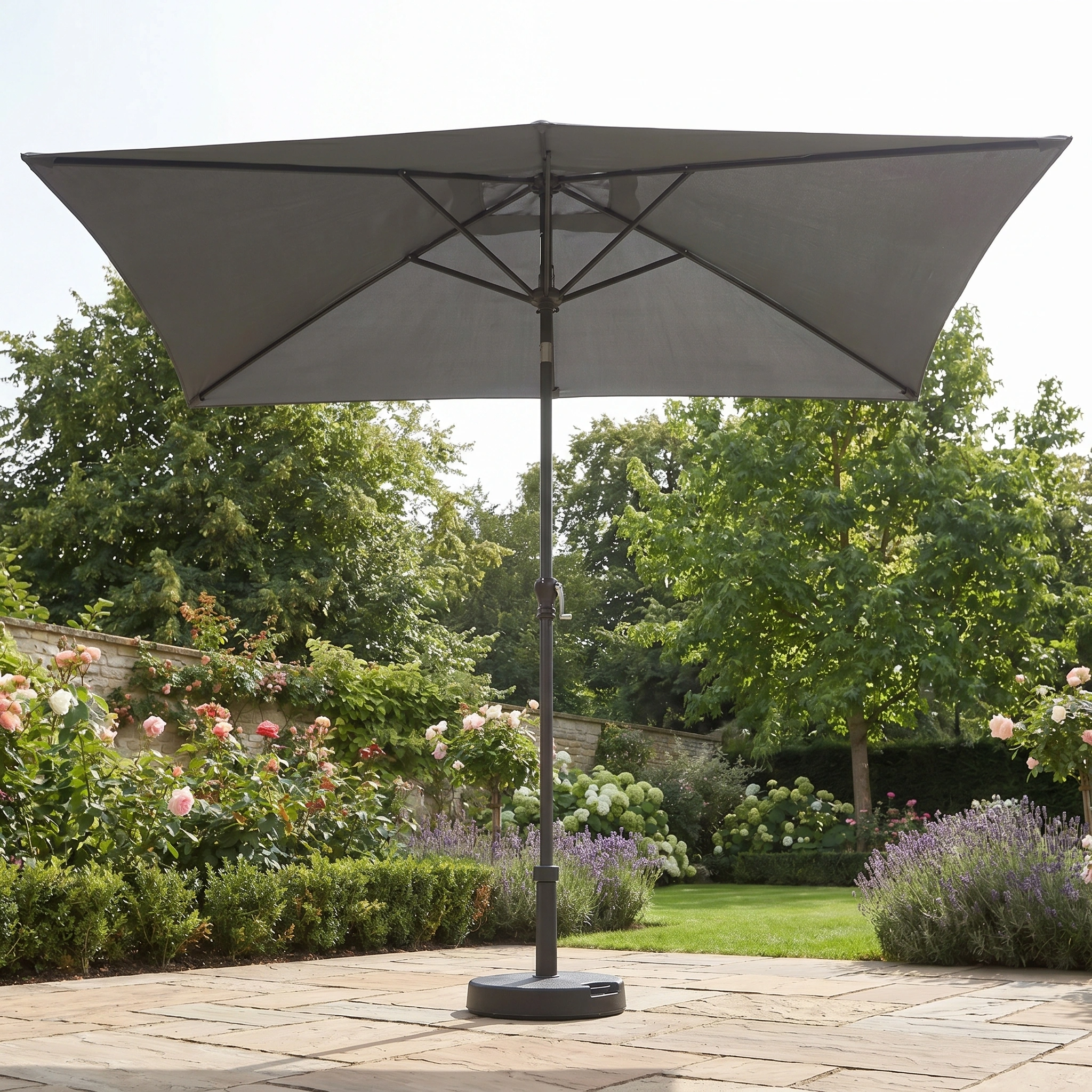 The Triton 3m x 2m Rectangular Aluminium Parasol in Grey stands open on a stone patio in a lush garden, offering UV sun protection with its durable aluminium frame.