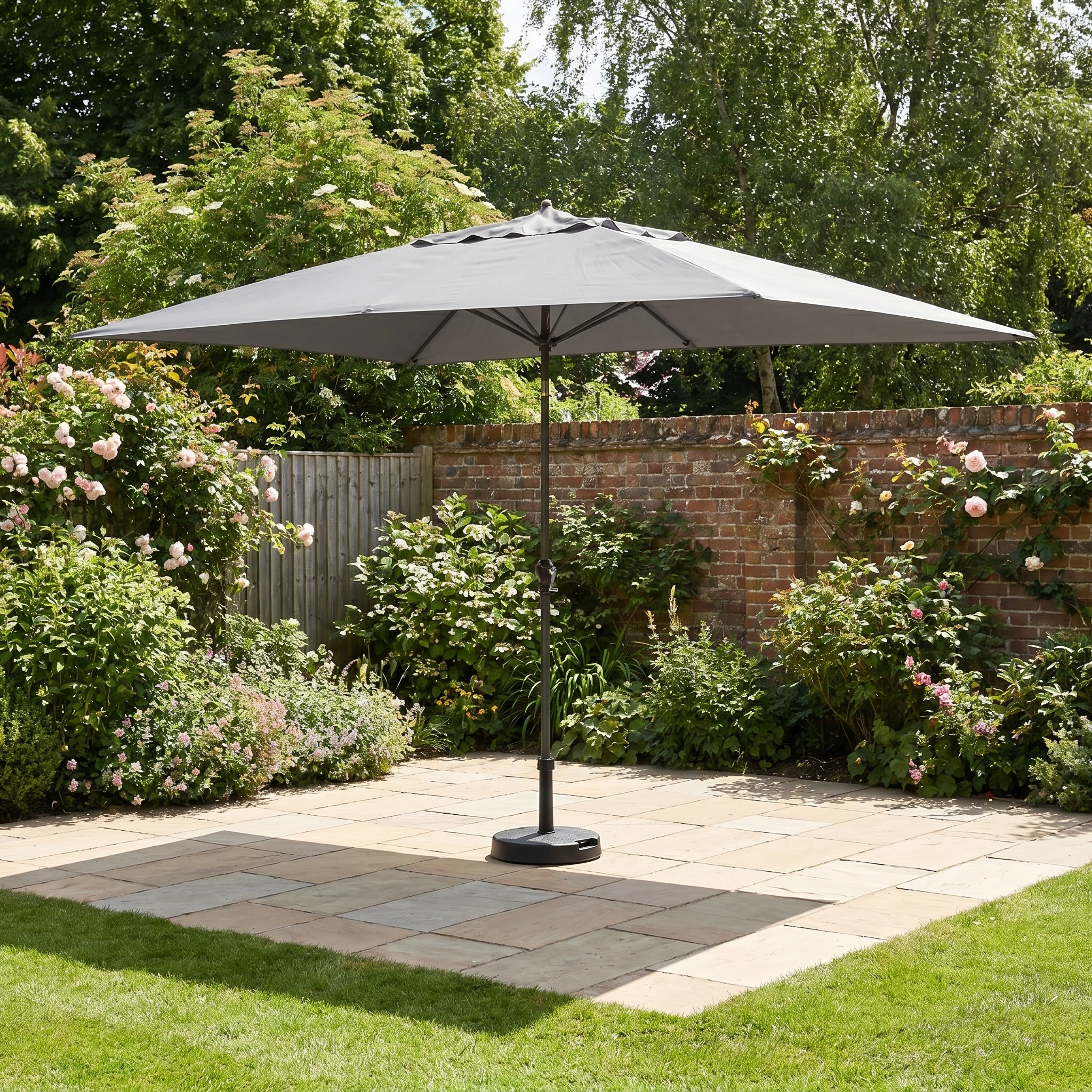 The Triton 3m x 2m Rectangular Aluminium Parasol in Grey offers stylish shade and UV protection on a paved area in a blooming, sunny garden.