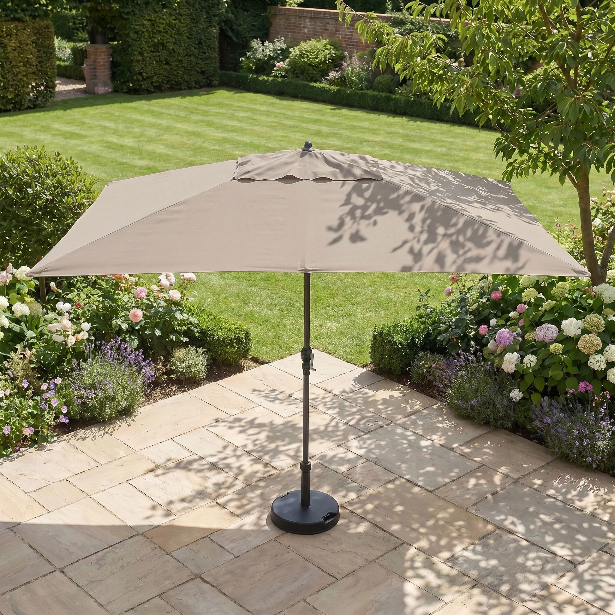 The Triton 3m x 2m Rectangular Aluminium Parasol in Khaki provides shade on a stone terrace, bordered by a lush green lawn and vibrant garden blooms.