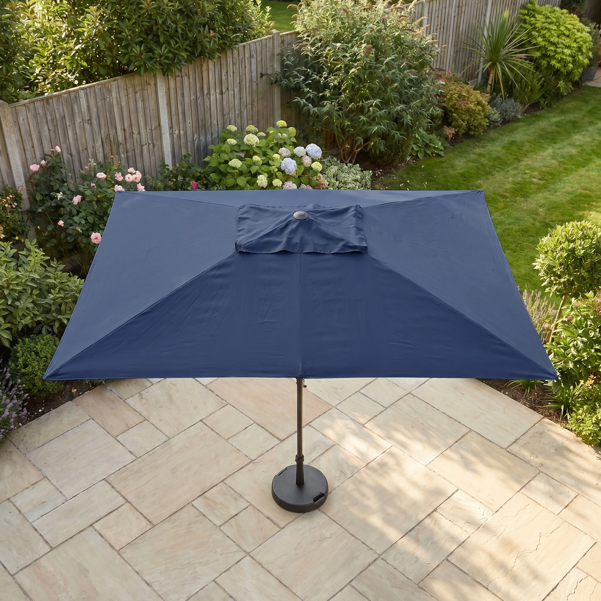 The Triton 3m x 2m Rectangular Aluminium Parasol in Navy, featuring UV protection, stands on a stone patio amidst a lush garden.