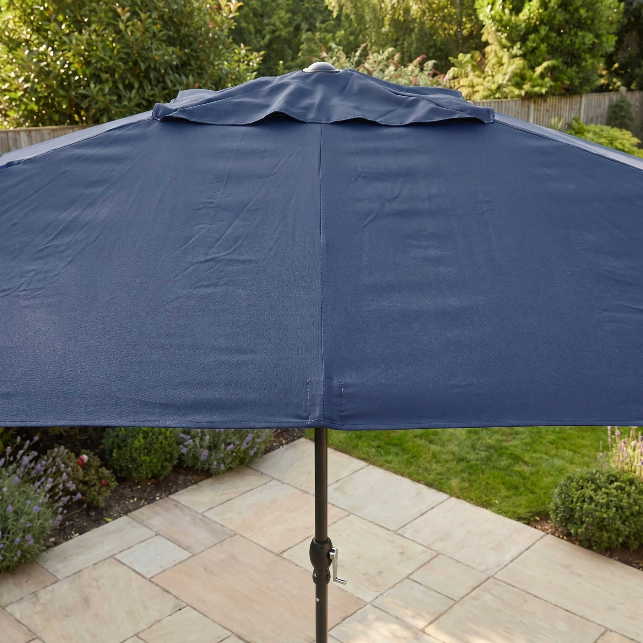 The Triton 3m x 2m Rectangular Aluminium Parasol in Navy provides UV protection and shade on a stone terrace, set amid lush garden greenery and plants.