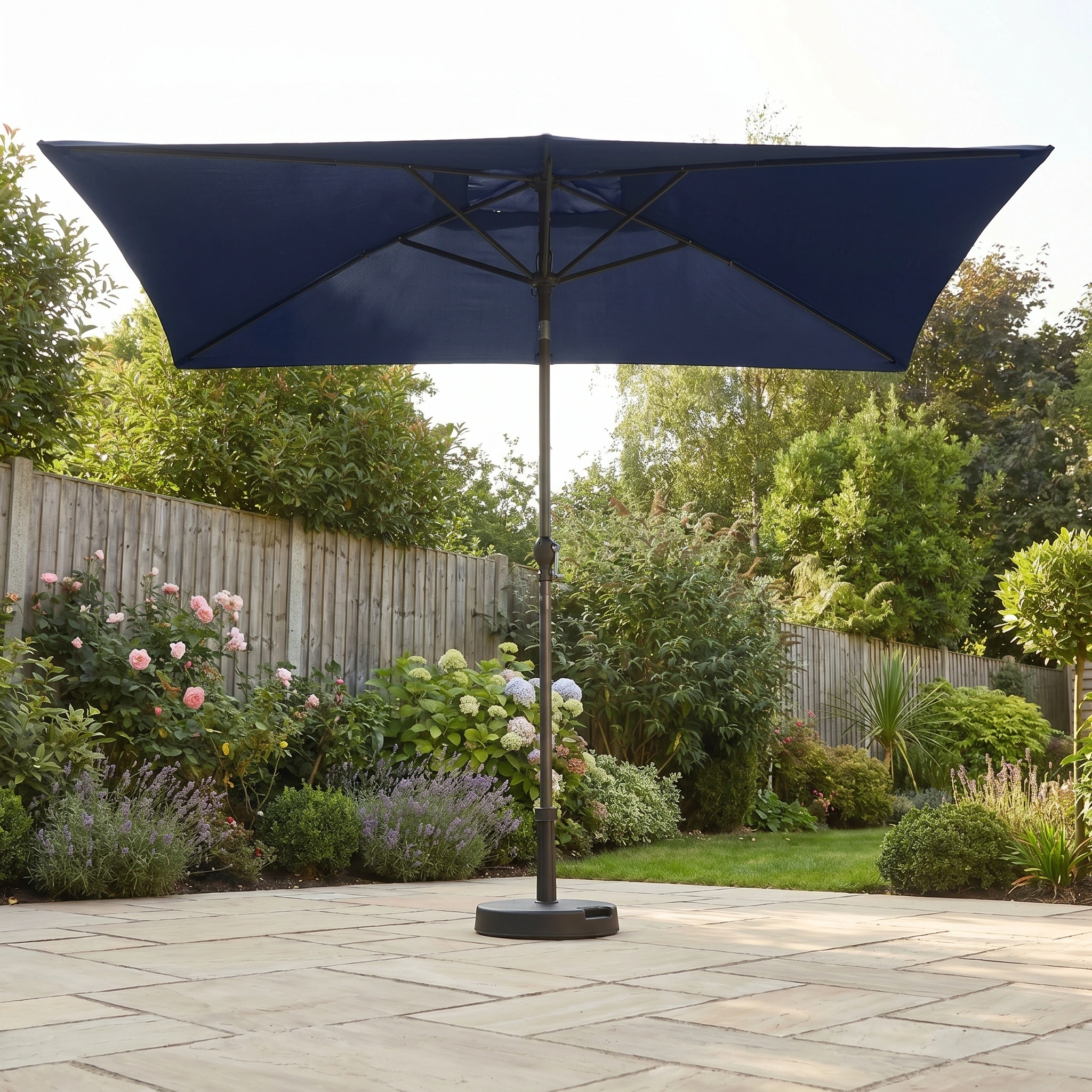 The Triton 3m x 2m Rectangular Aluminium Parasol in Navy stands on a stone terrace in a flower-filled garden, providing UV protection amidst greenery.