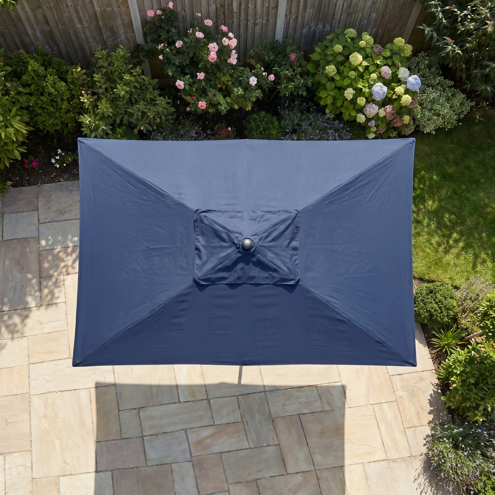 The Triton 3m x 2m Rectangular Aluminium Parasol in Navy with UV protection shades a stone patio surrounded by flowers and green plants.