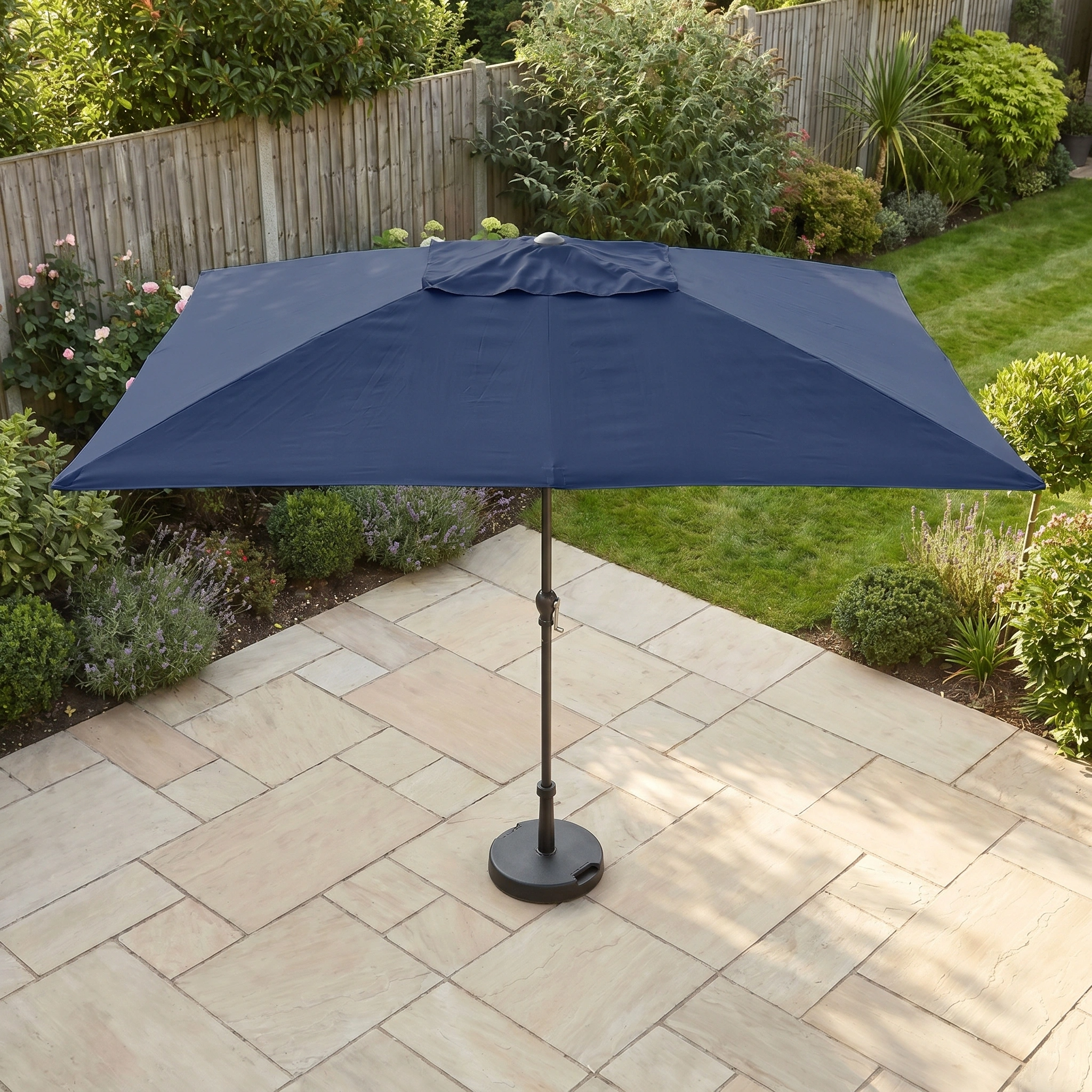 The Triton 3m x 2m Rectangular Aluminium Parasol in Navy stands on a stone patio in a garden with green plants and a wooden fence, offering superior UV protection.