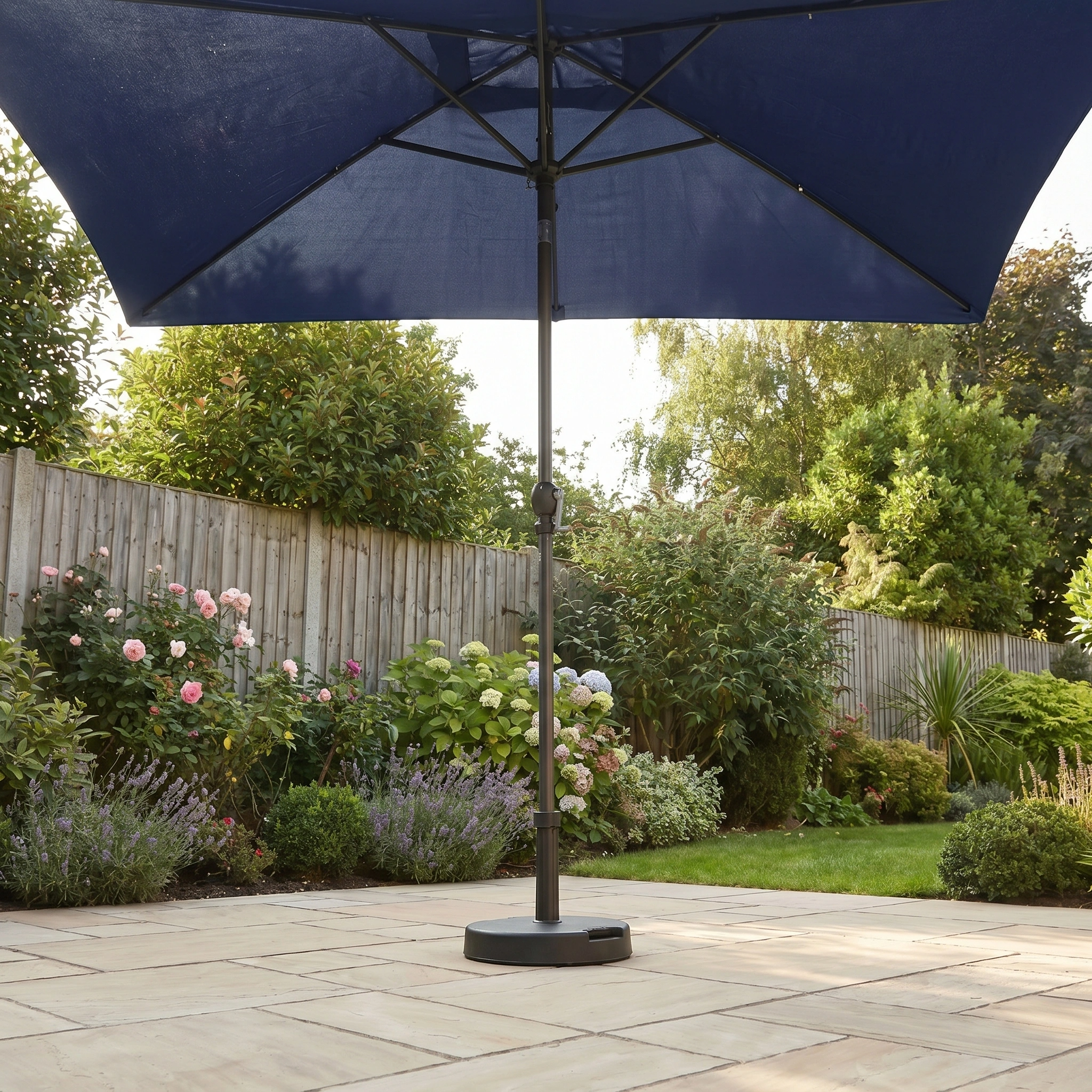 The Triton 3m x 2m Rectangular Aluminium Parasol in Navy stands on a sunny patio with flowering plants and a wooden fence, providing excellent UV protection.