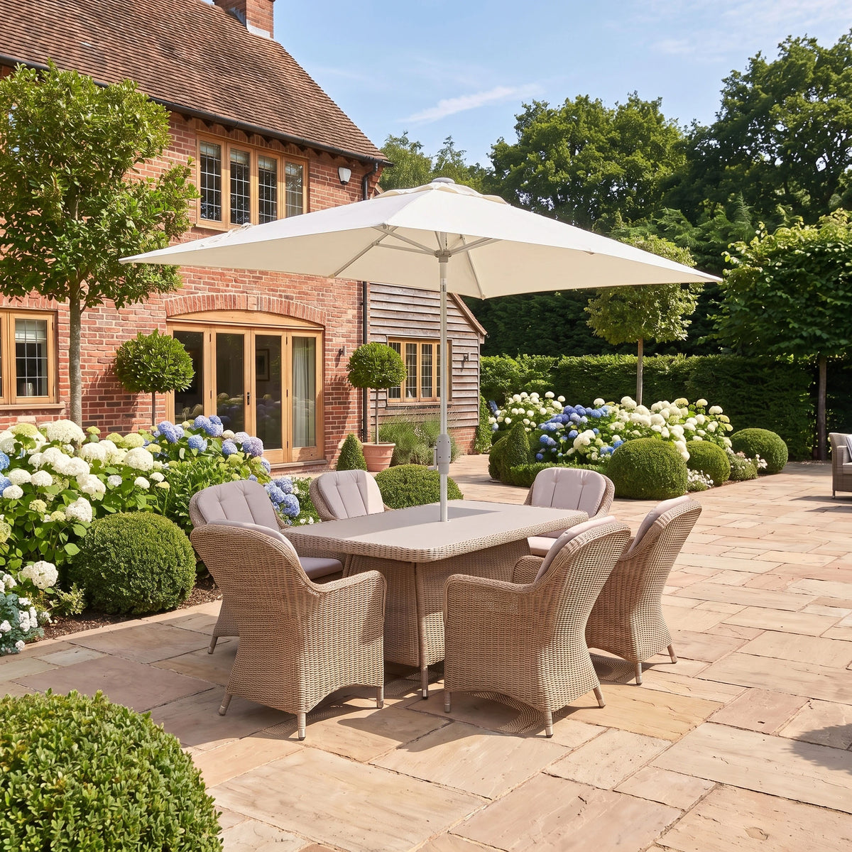 Outdoor patio featuring the Vela 6 Seat Rectangular Dining Set in Wheat with a rectangular table and umbrella, surrounded by neatly trimmed bushes beside a brick house.